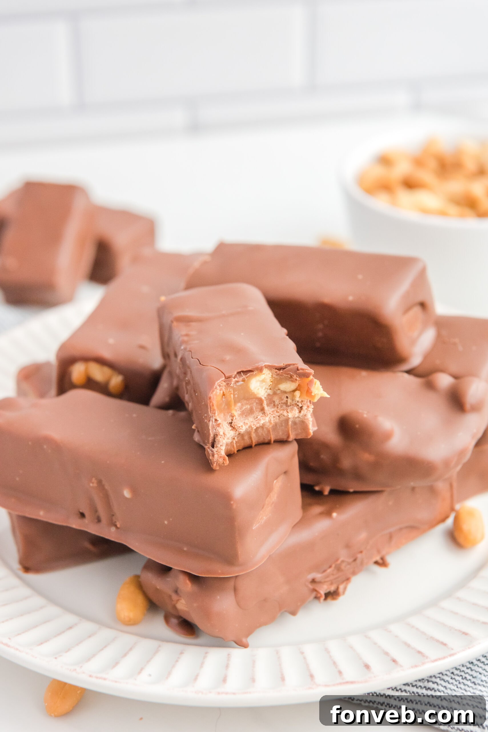 Homemade Snickers Candy Bars piled on a white serving plate with a bite missing from one, showcasing the intricate layers of nougat, caramel, and peanuts under a rich chocolate coating.