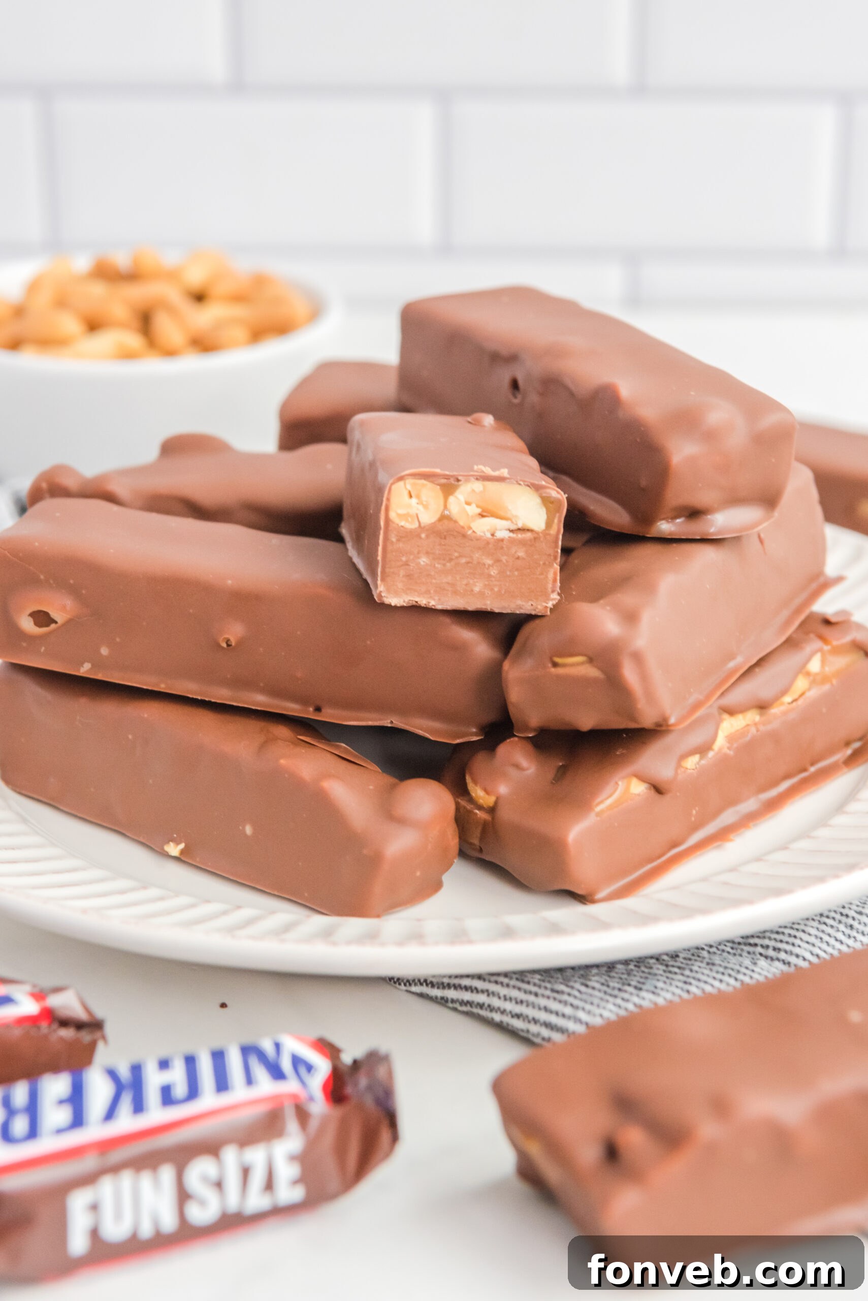 Front view of Homemade Snickers Bars stacked on a white plate with one bar expertly cut in half to reveal the rich interior layers.