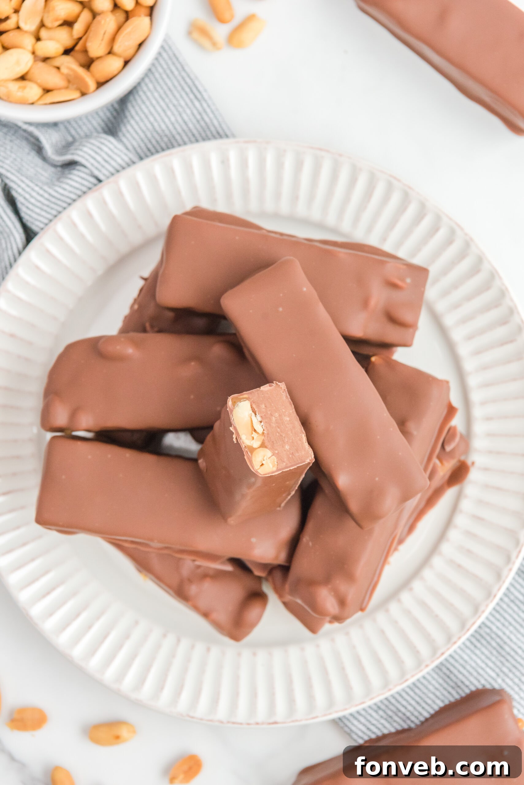 Overhead view of Homemade Snickers Bars stacked on a white plate with one bar cut in half, revealing the distinct layers of chocolate, caramel, peanuts, and nougat.