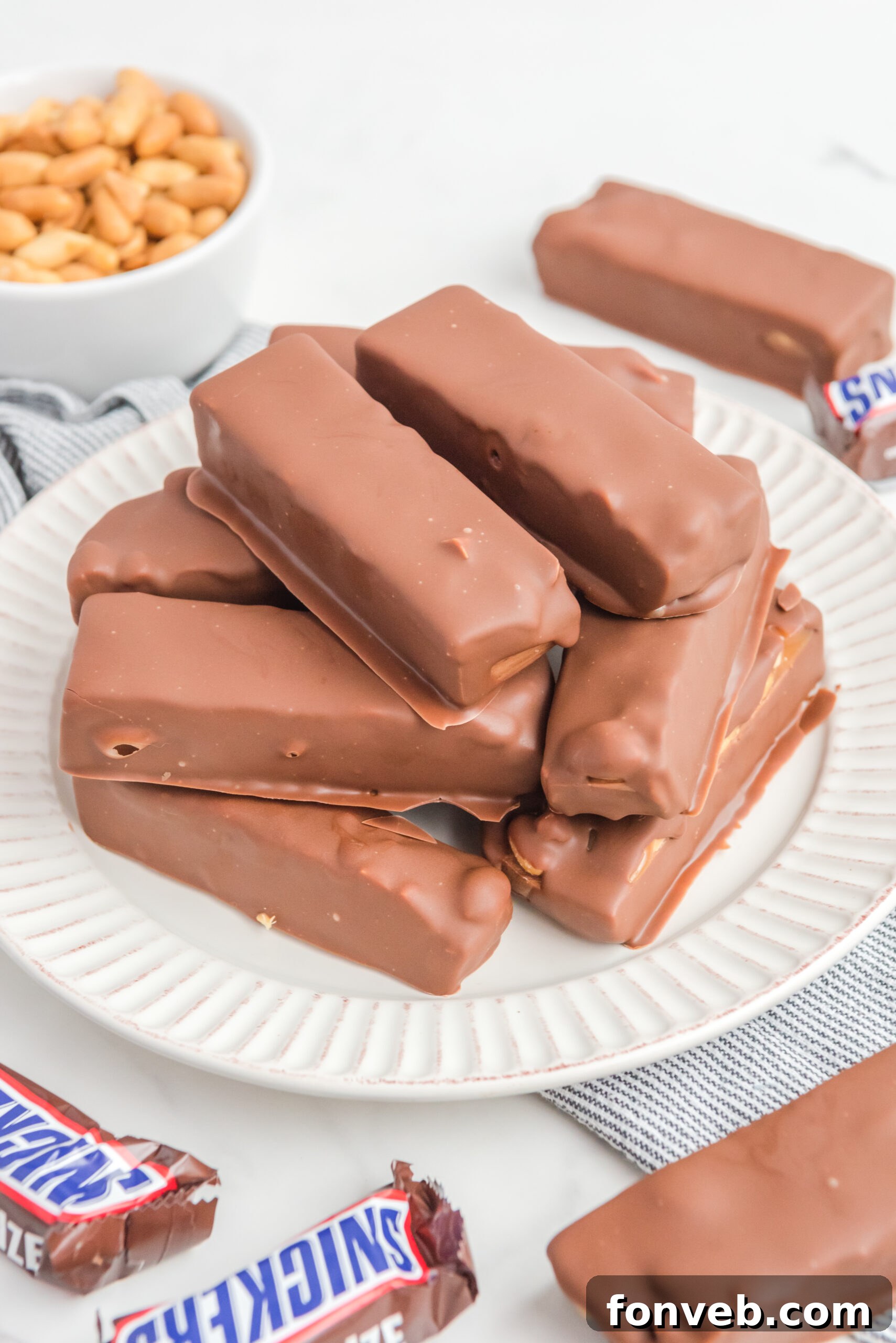 Homemade Snickers Candy Bars piled on a white serving plate, gleaming under their chocolate coating.