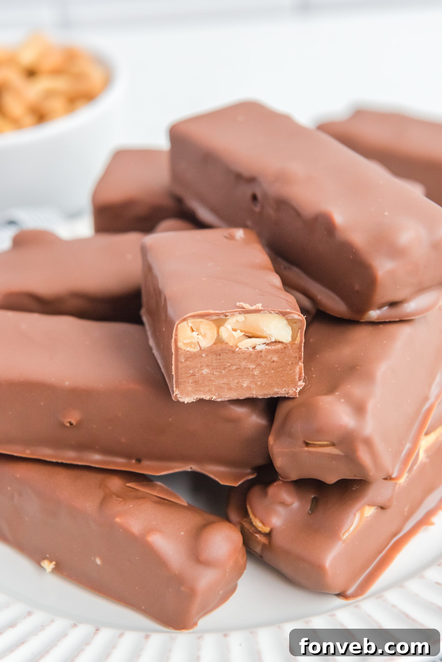 Homemade Snickers Candy Bars piled on a white serving plate with one cut open to show the inside, clearly displaying the nougat, peanuts, and caramel layers.