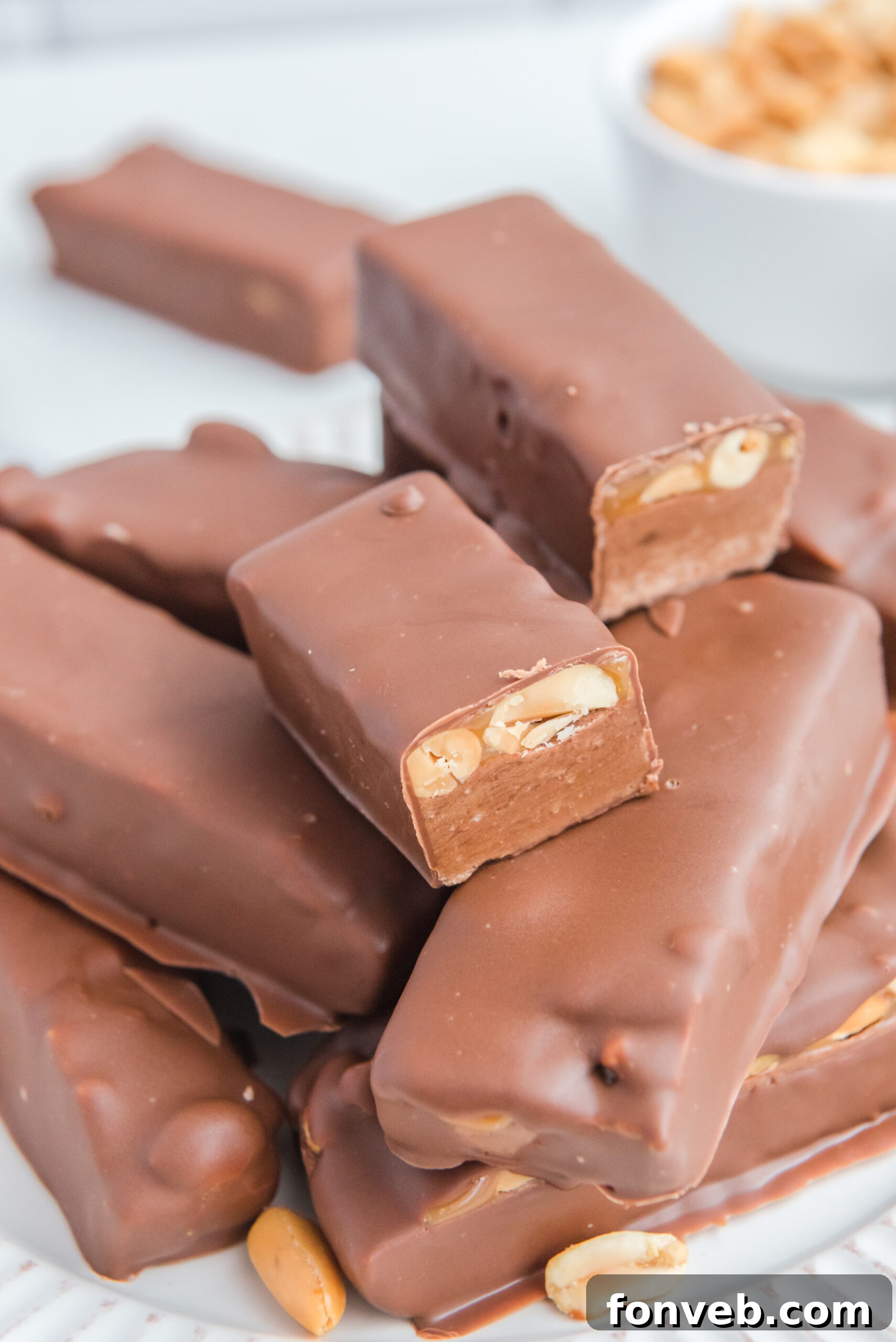 Homemade Snickers Candy Bars piled high on a white serving plate with a bite missing from one, enticingly showing off its interior.