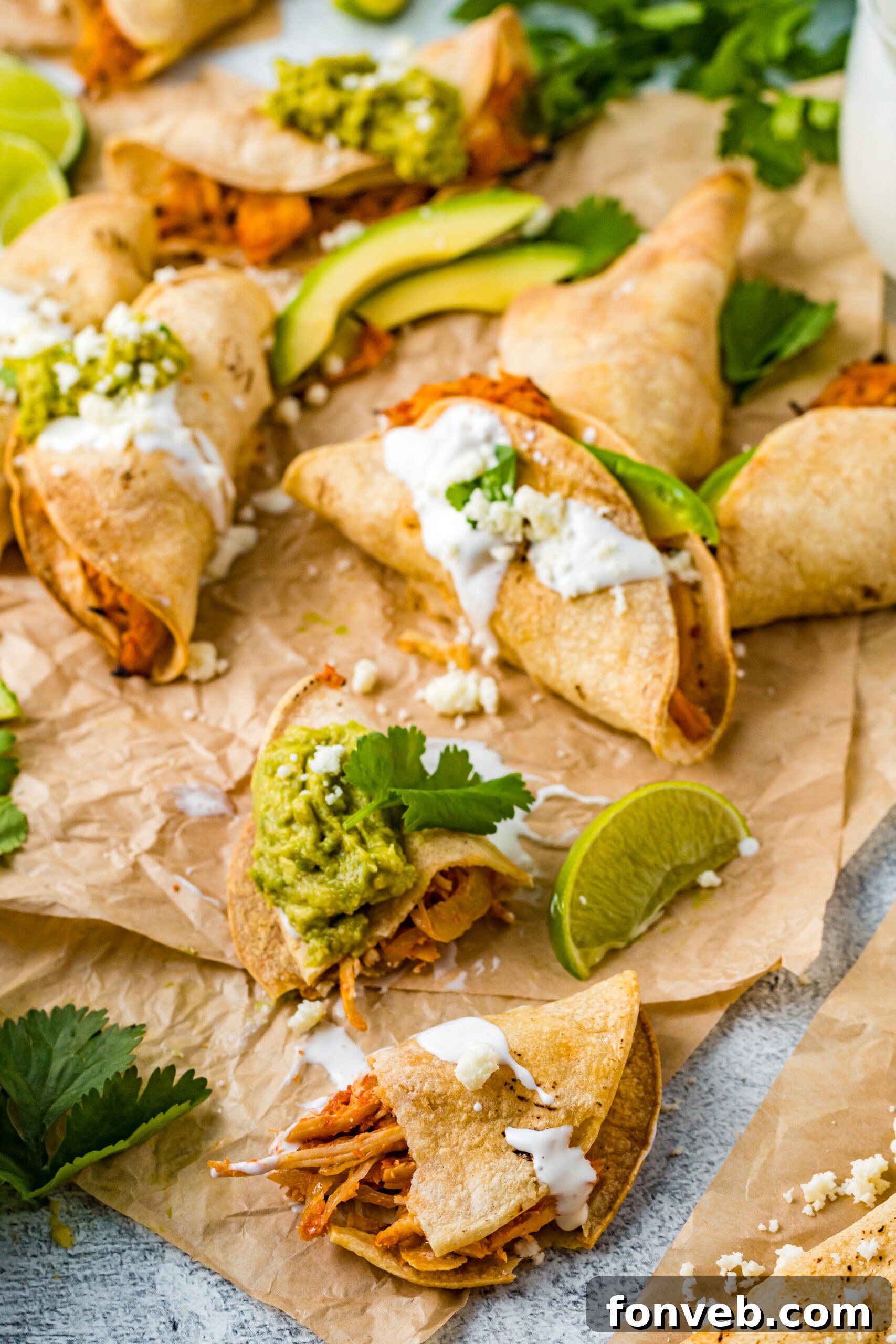 Front view of Crispy Tinga Tacos on a piece of parchment paper surrounded by lime and avocado slices with one taco split in half.