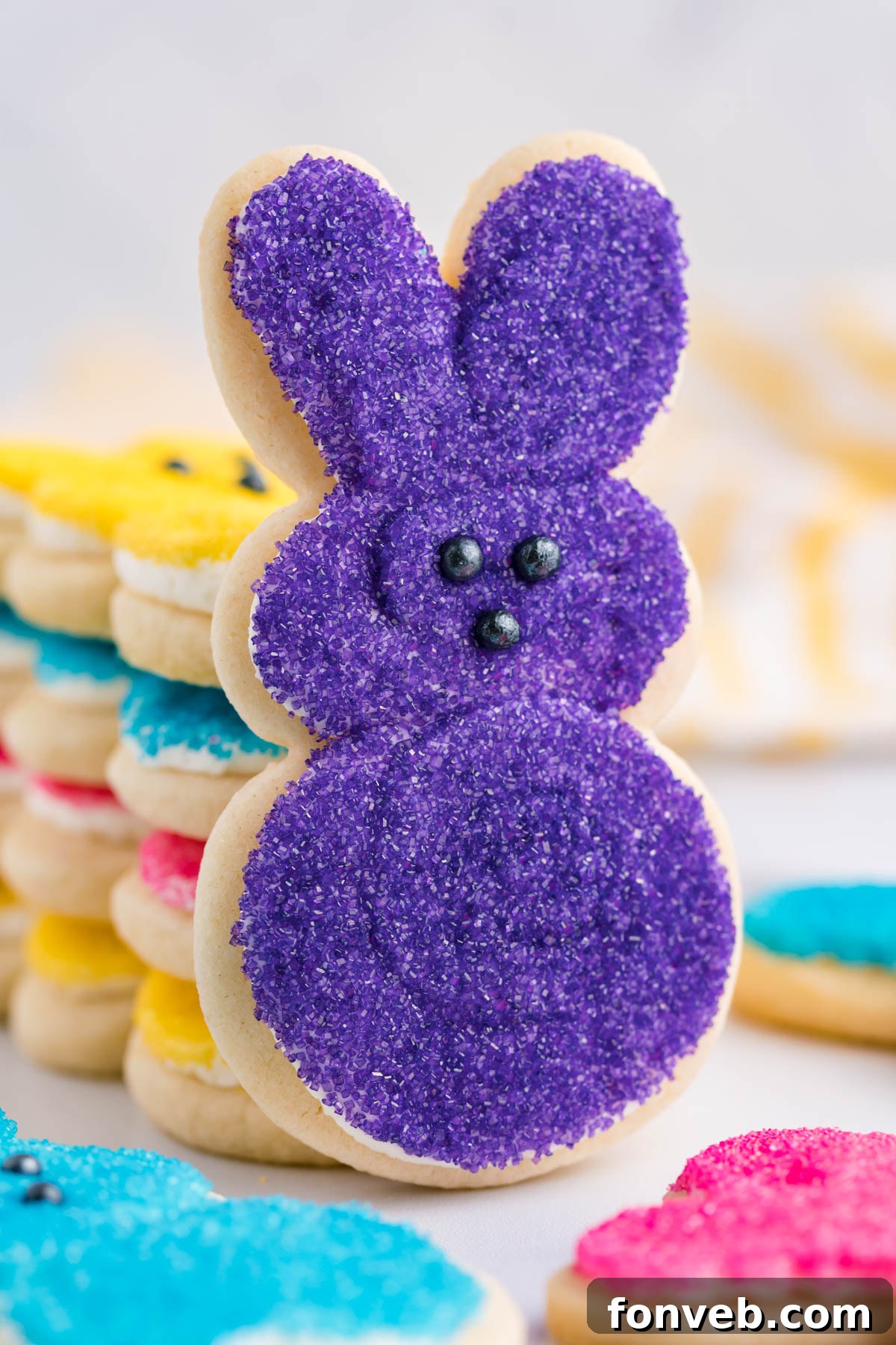 A beautifully frosted purple Bunny Cookie artfully propped up against a stack of multicolored Easter Bunny Cookies, highlighting their varied hues and charming appeal.