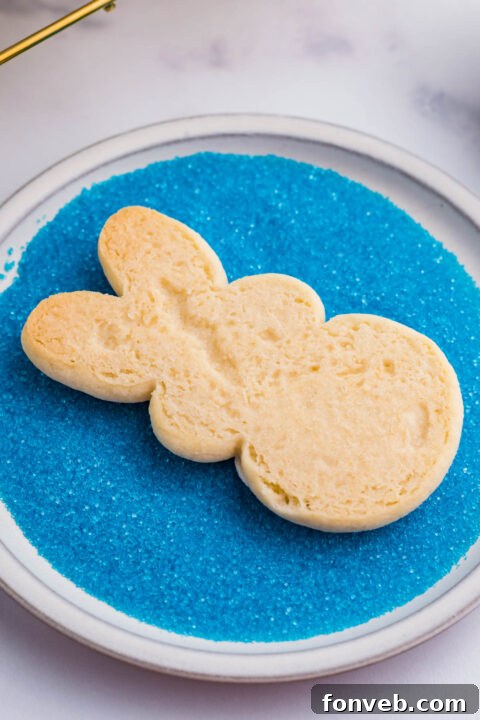 A bunny cookie being carefully dipped into a bowl of blue sprinkles, ensuring a vibrant, even coating.