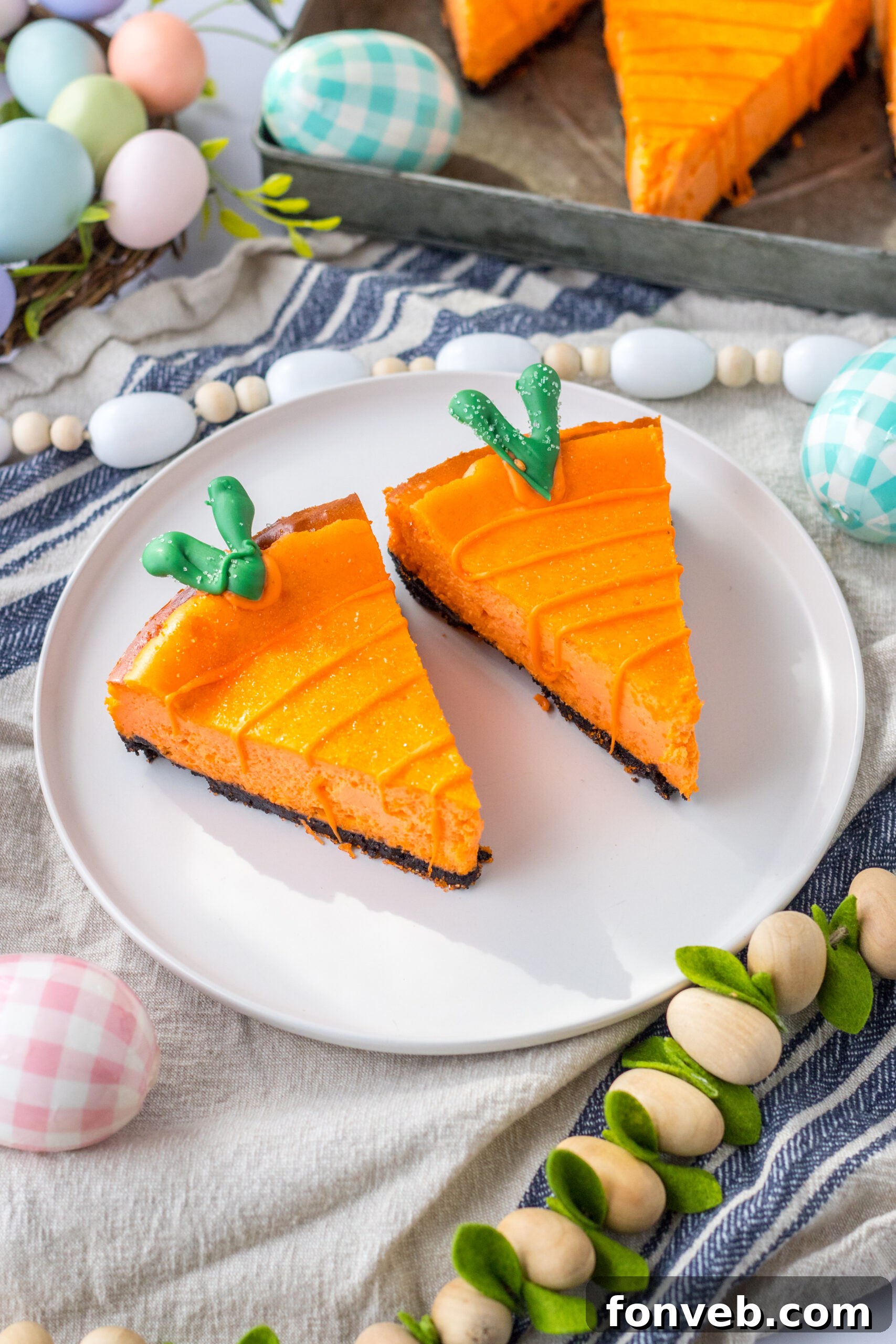 Two delightful cheesecake carrots are artfully arranged on a white plate, set against a soft, blurred background of white and blue linen, enhancing their festive appeal.