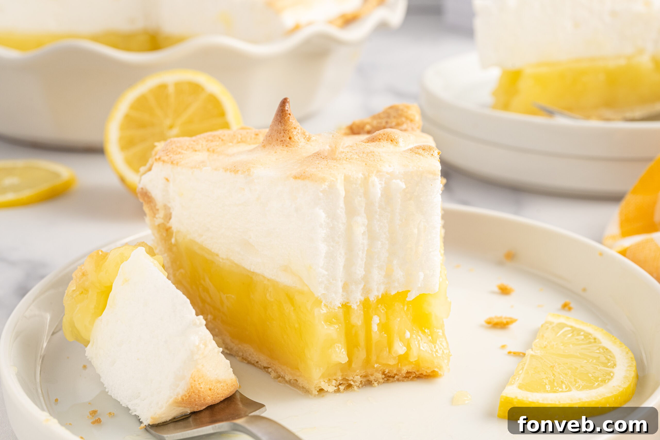 up close view of a piece of the lemon meringue pie with a bite missing on a white plate with fresh sliced lemons. 