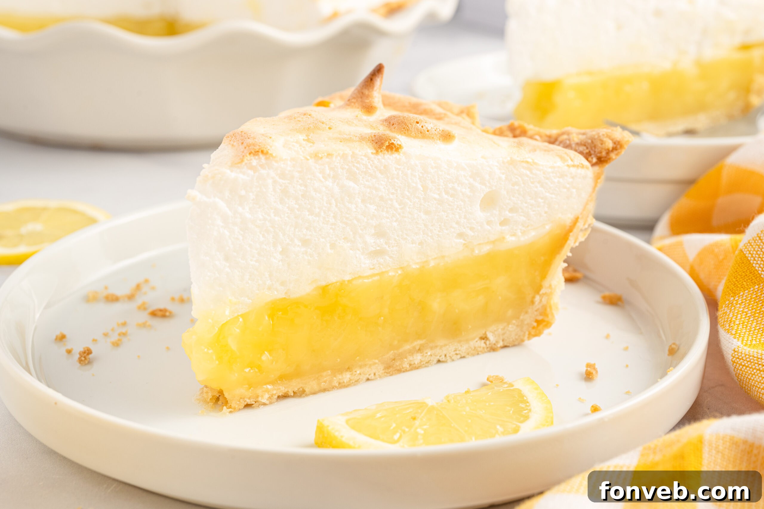 side view of a piece of the lemon meringue pie on a white plate with fresh sliced lemons. 