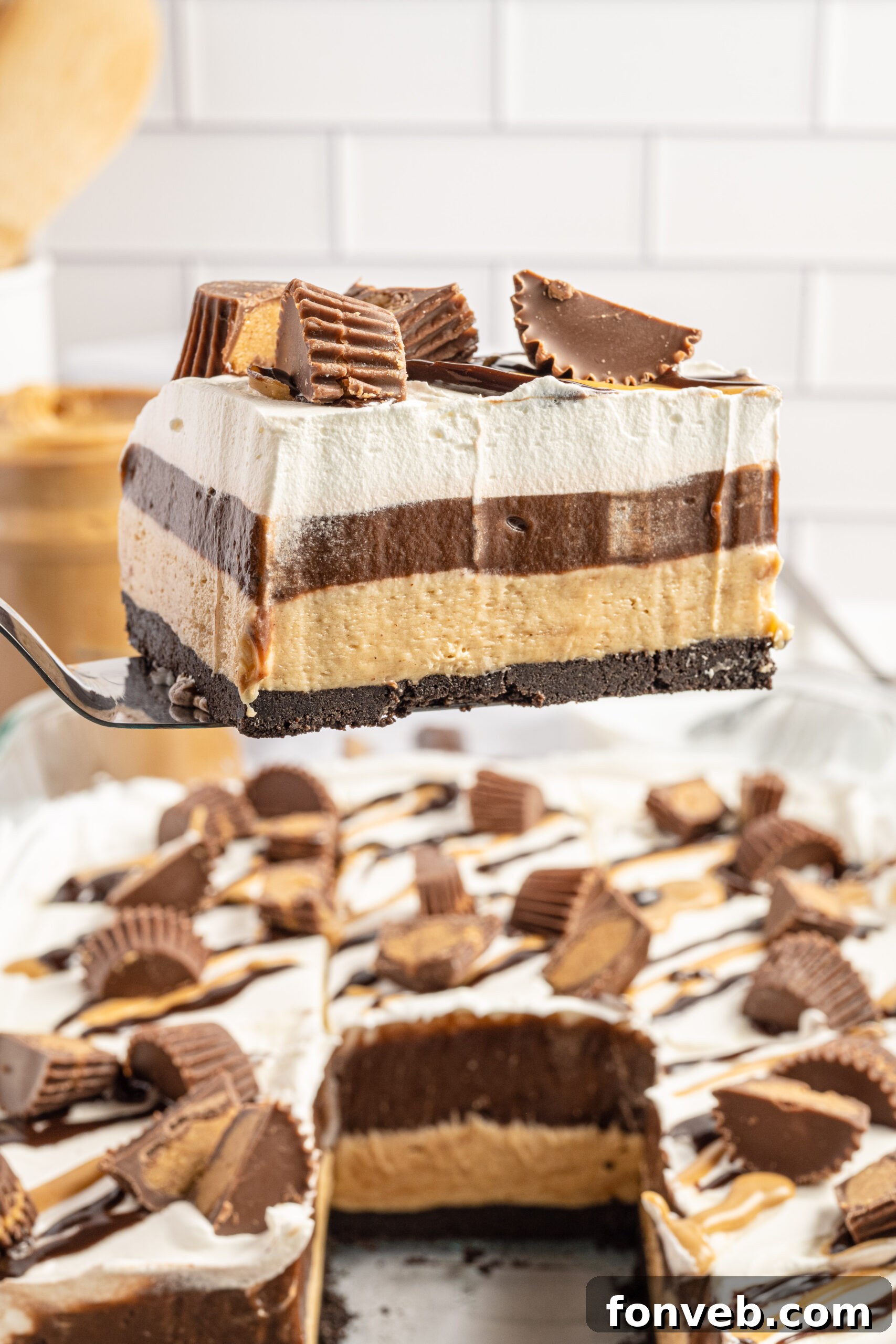 Reese's Peanut Butter Cup Lasagna 2 A slice of Reese's chocolate peanut butter cup lasagna being carefully lifted from a glass baking dish with a silver spatula, revealing its distinct creamy layers.