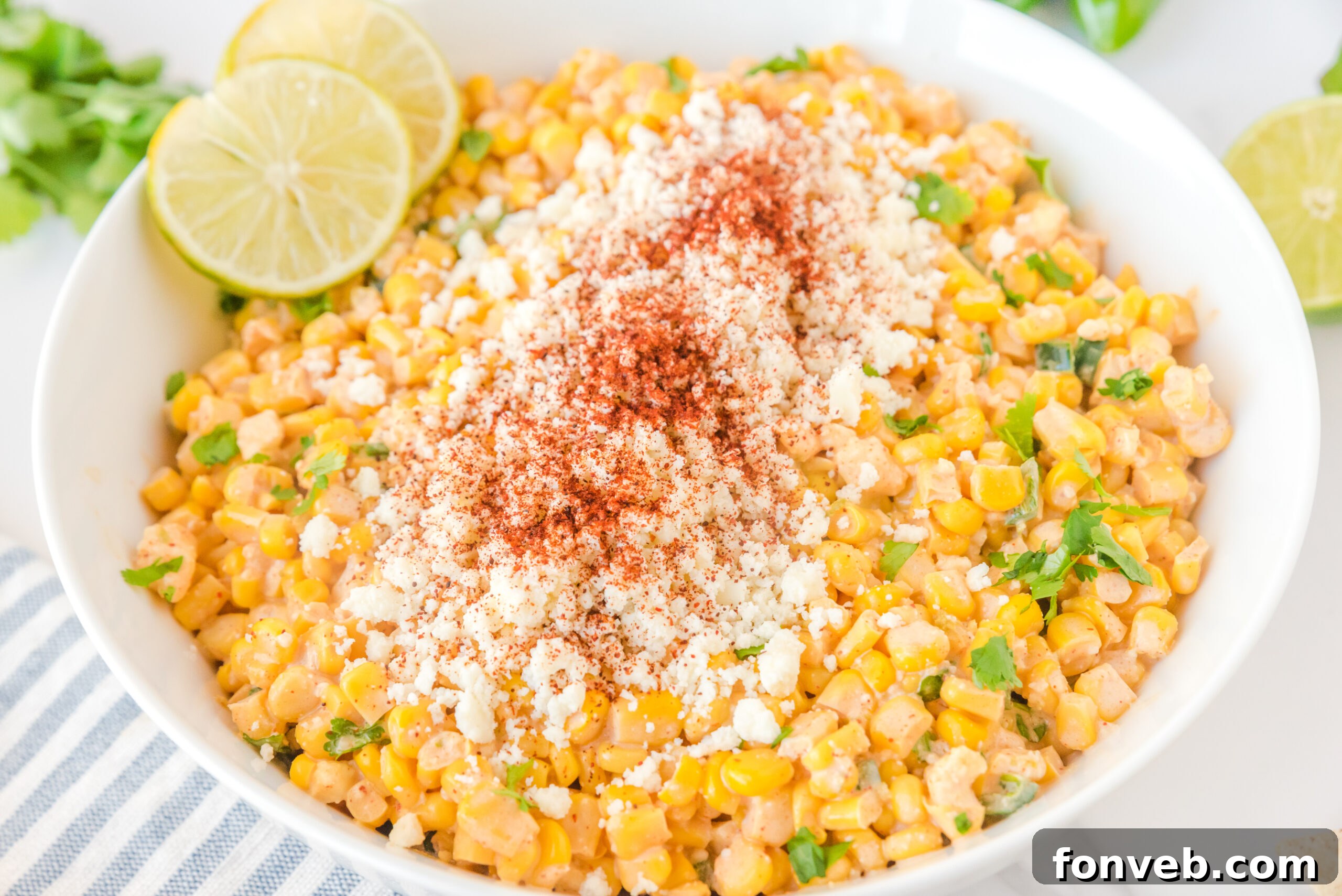 A generously sized white serving bowl filled with Mexican Street Corn Salad, elegantly topped with crumbled feta cheese and an array of fresh lime slices.