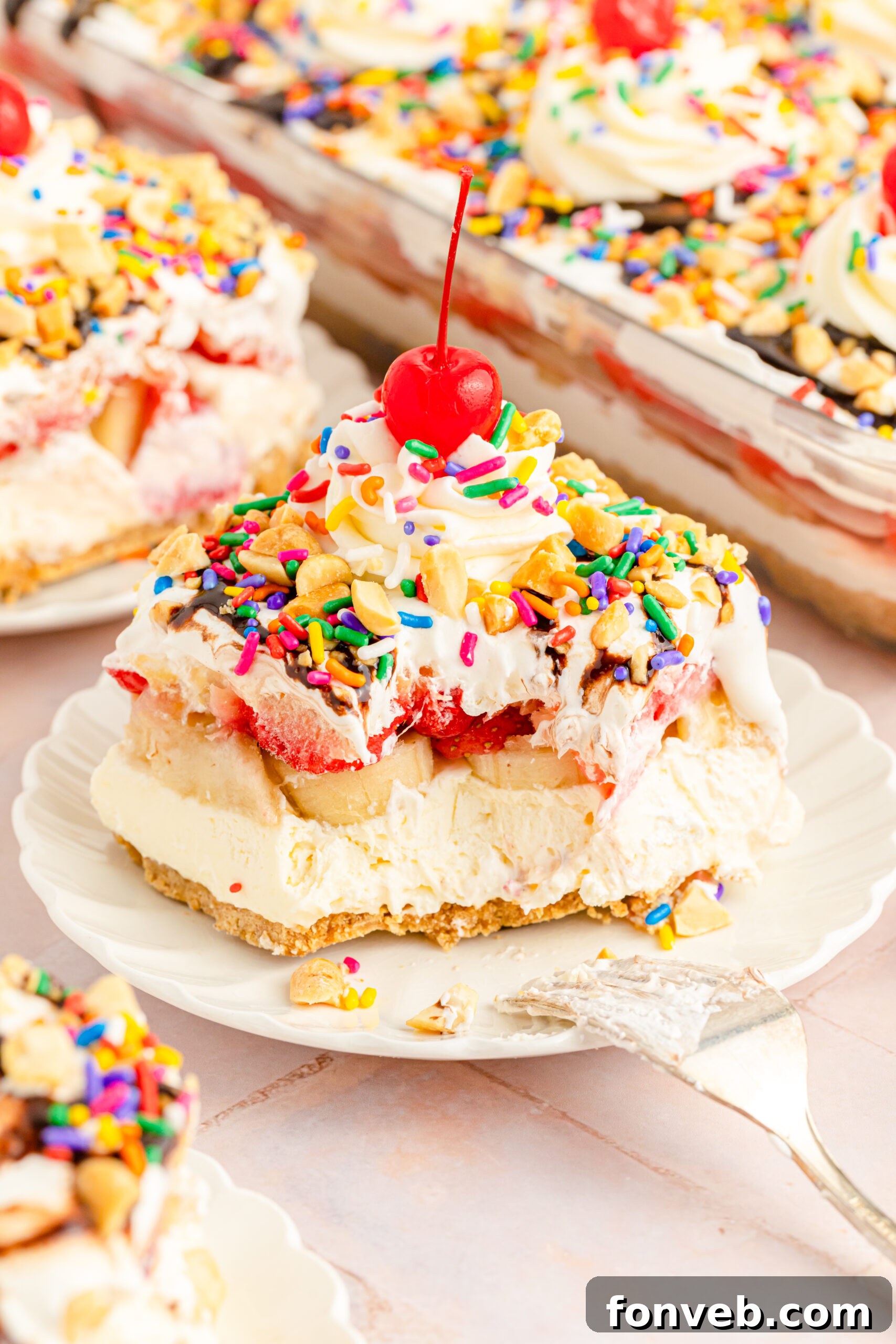 A front perspective of a slice of No-Bake Banana Split Cake on a white plate, featuring a gentle bite removed by a silver fork, inviting indulgence.