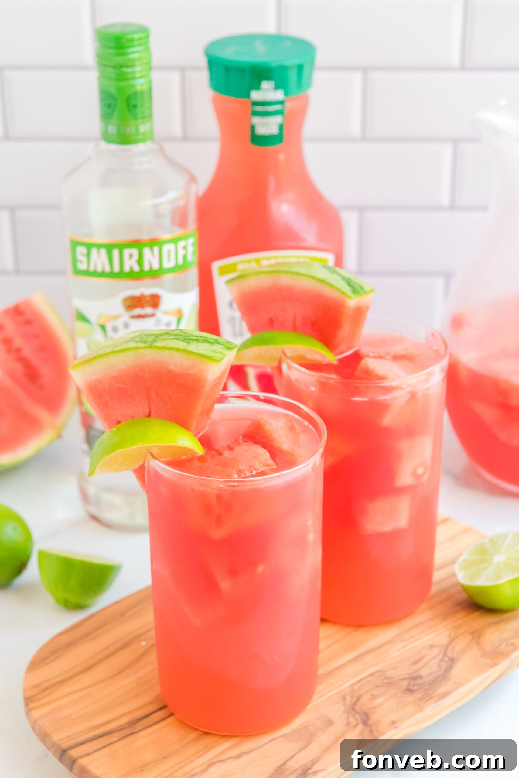 Front view of Watermelon Cocktail in two clear glasses garnished with lime and watermelon wedges on a wooden board.