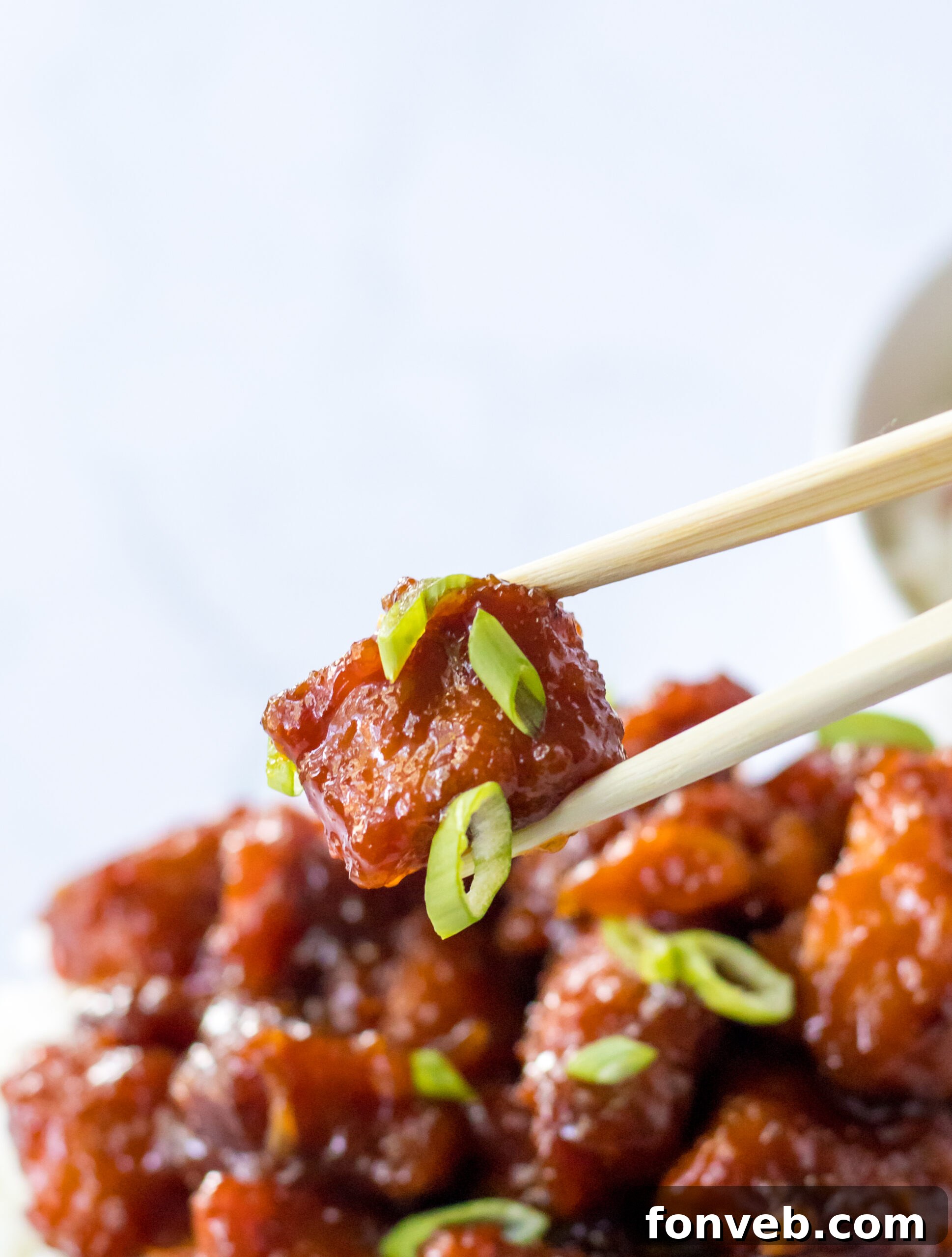 Close up view of chopsticks holding a piece of Slow Cooker Firecracker Chicken, beautifully plated with rice and scallions.