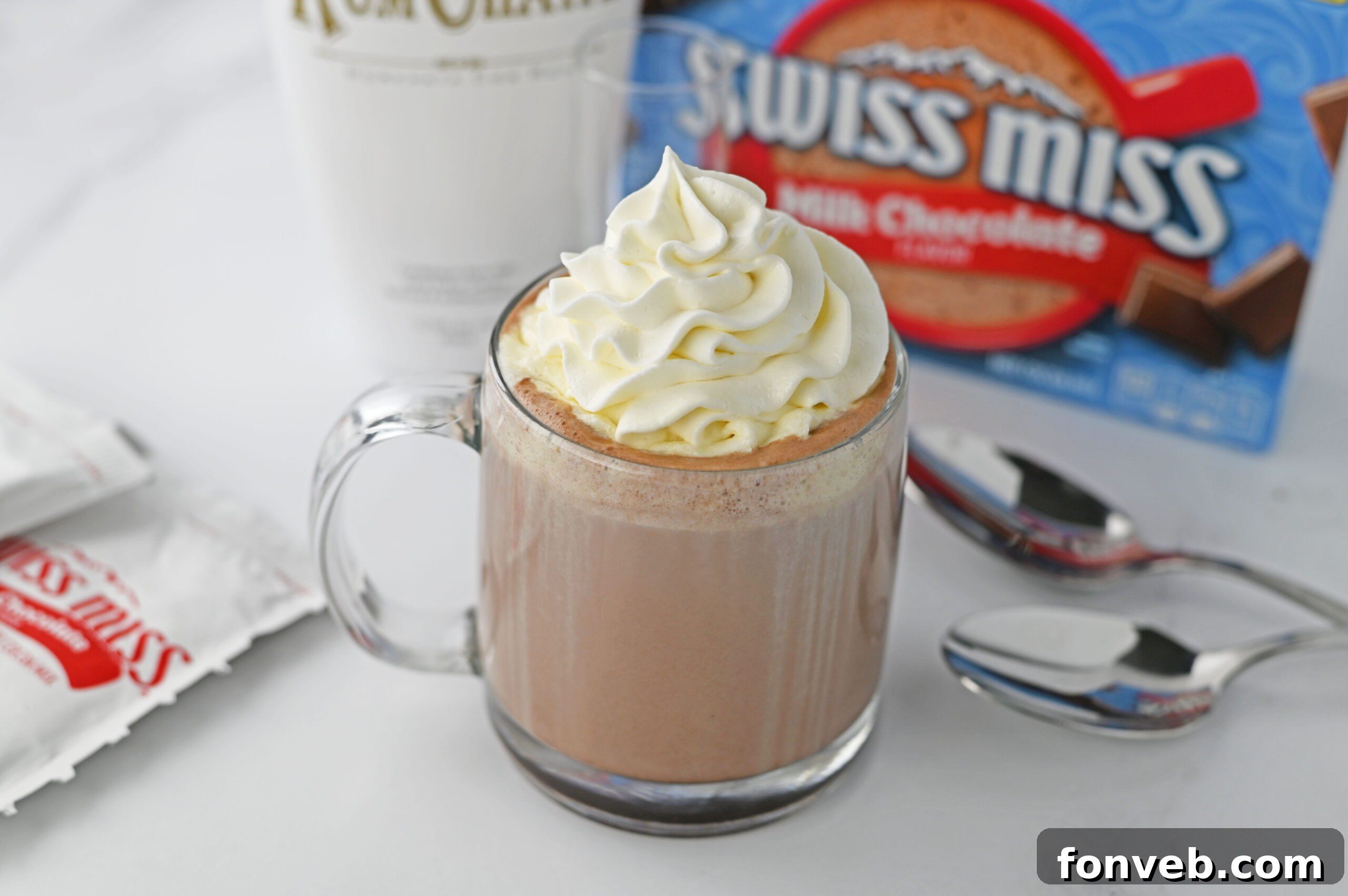 A clear glass mug of RumChata hot chocolate with whipped cream, positioned beside a classic Swiss Miss hot chocolate box and a bottle of RumChata liqueur