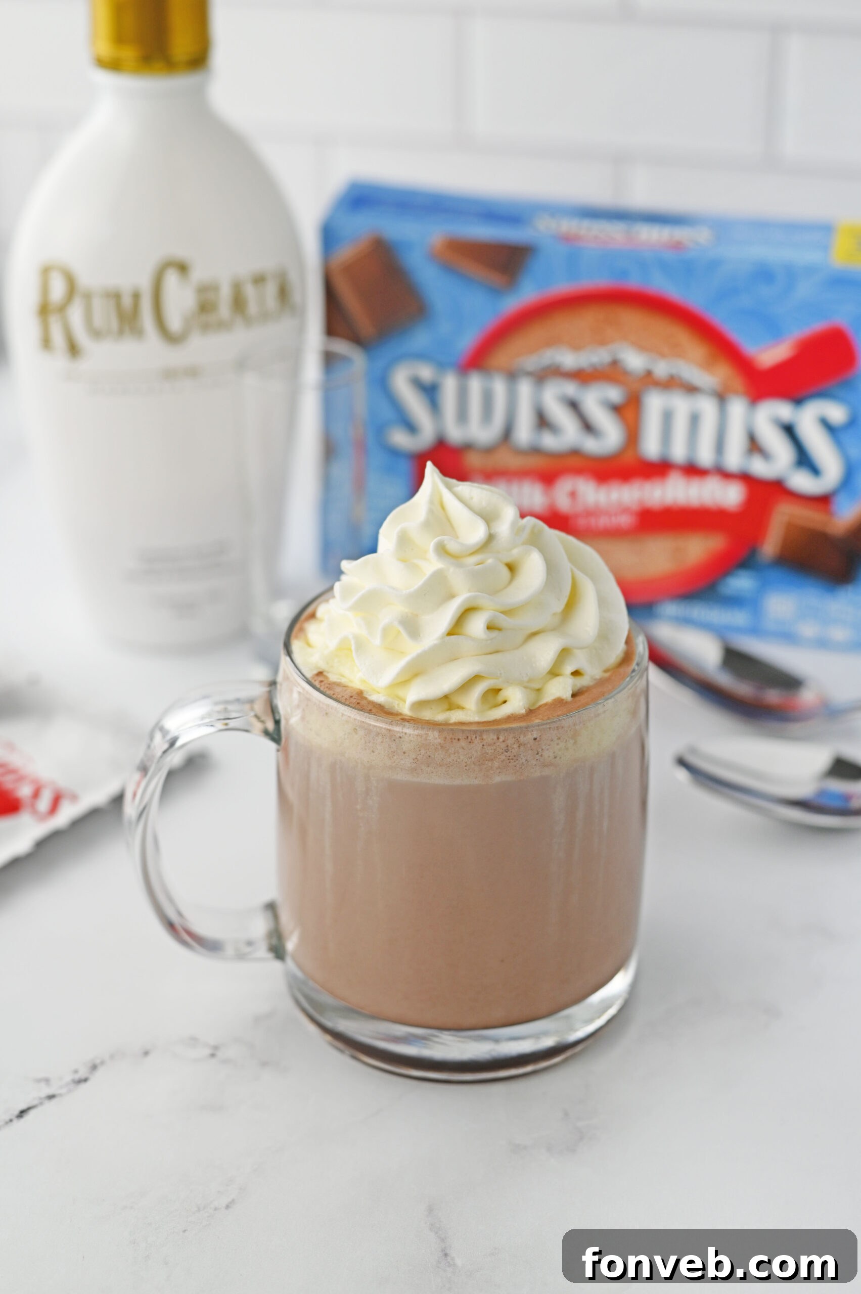 A clear glass mug of RumChata hot chocolate, generously topped with whipped cream, resting on a white marble table