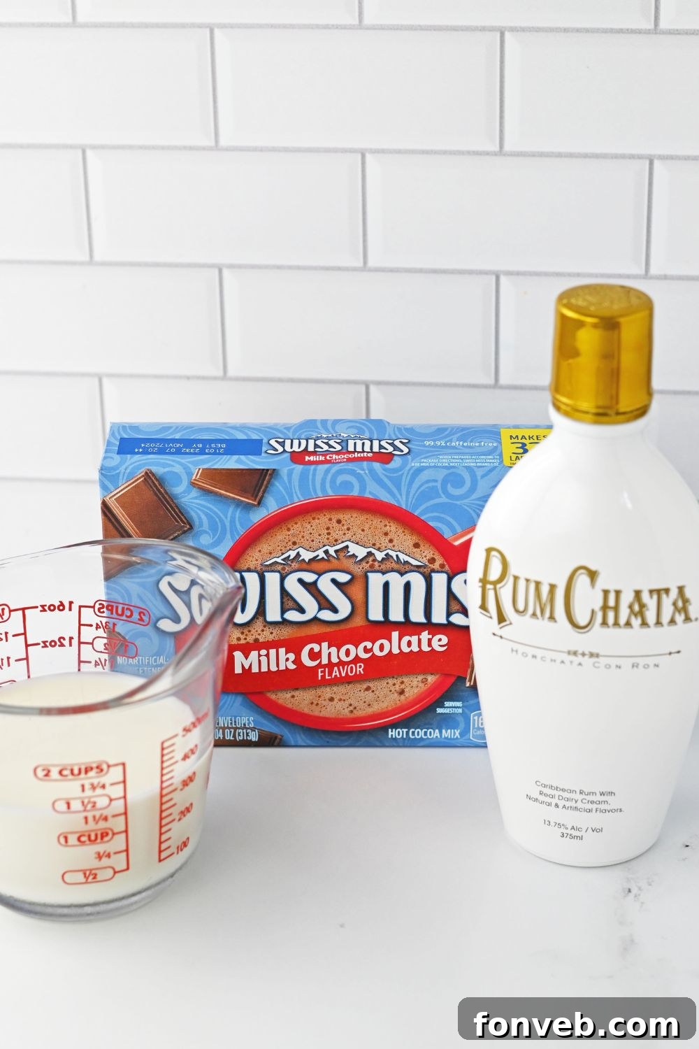 Ingredients for RumChata hot chocolate laid out on a white table: a packet of hot chocolate mix, a carton of milk, a bottle of RumChata, and a can of whipped cream