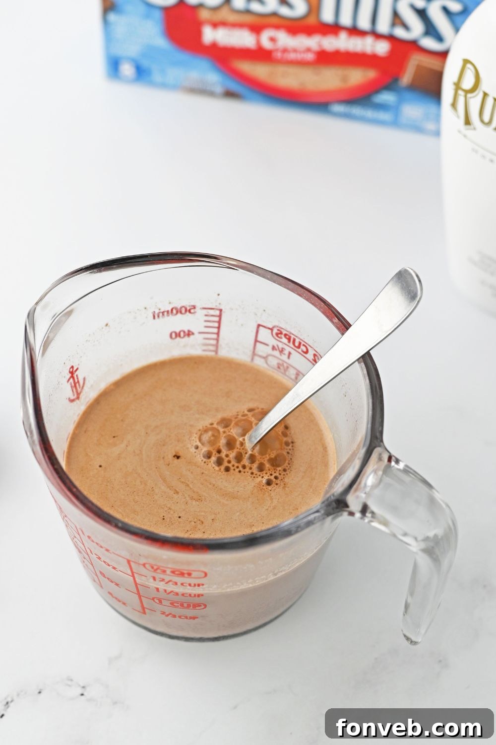 Mixing hot chocolate powder into warm milk in a clear measuring cup on a kitchen table