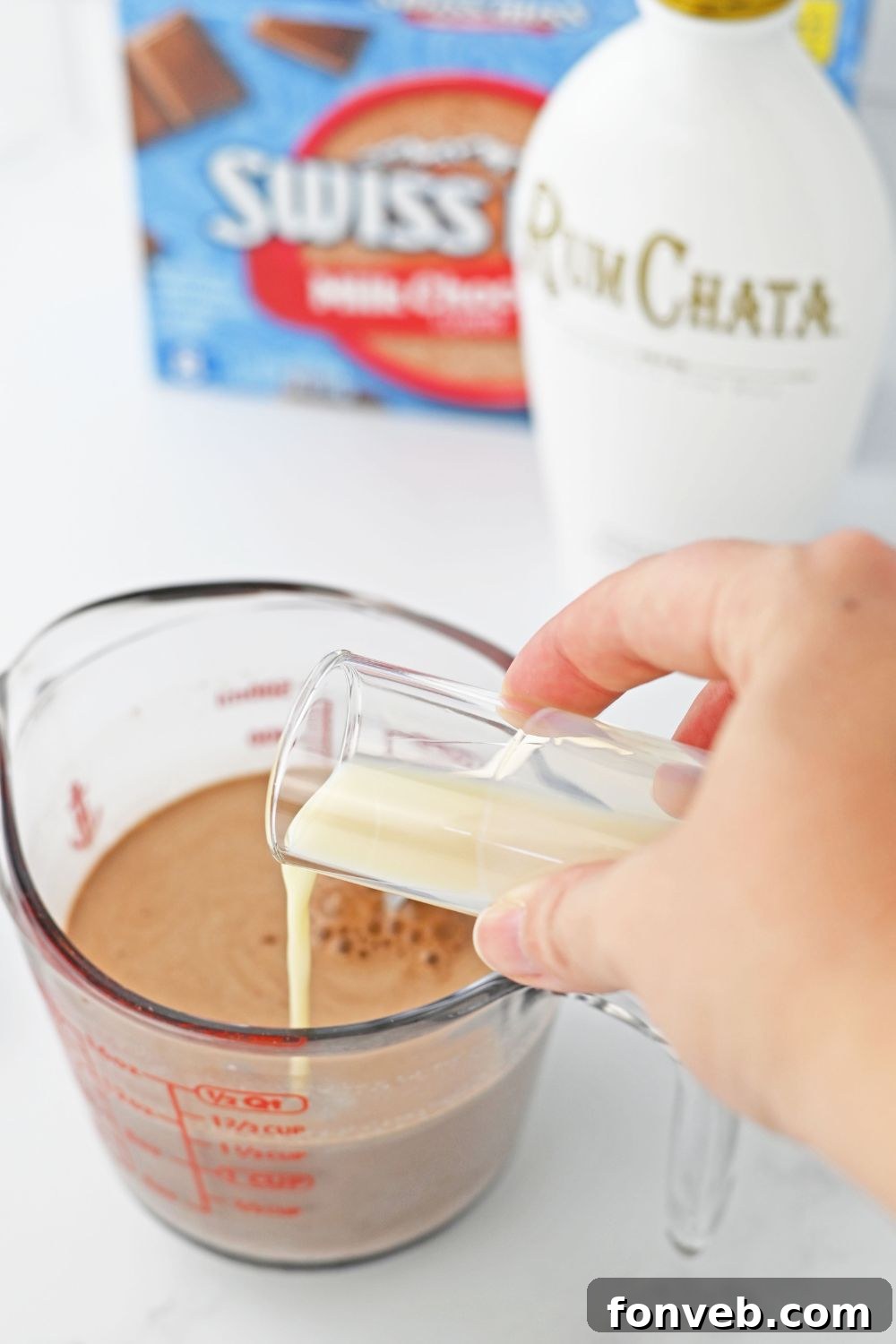 Pouring RumChata liqueur from a shot glass into a clear measuring cup containing mixed hot chocolate