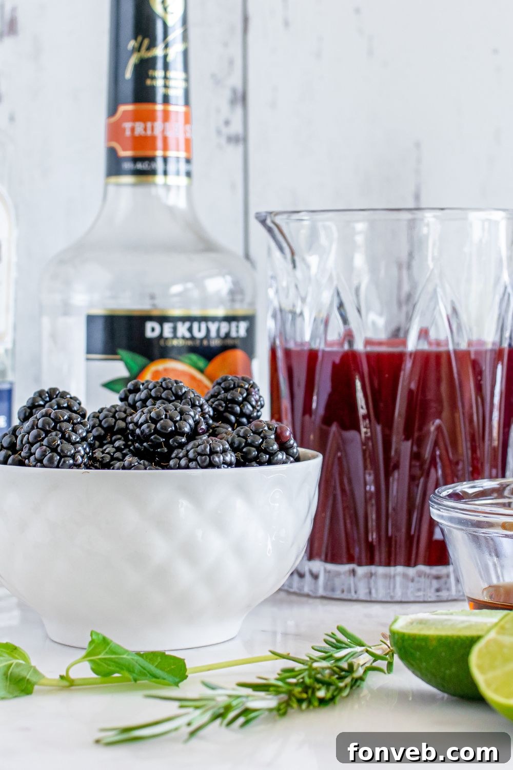 Magic Potion Margarita 6 An array of fresh ingredients for the Sanderson Sisters Margarita: blackberries in a bowl, a glass of vibrant juice, sliced limes, and other garnishes on a wooden table.