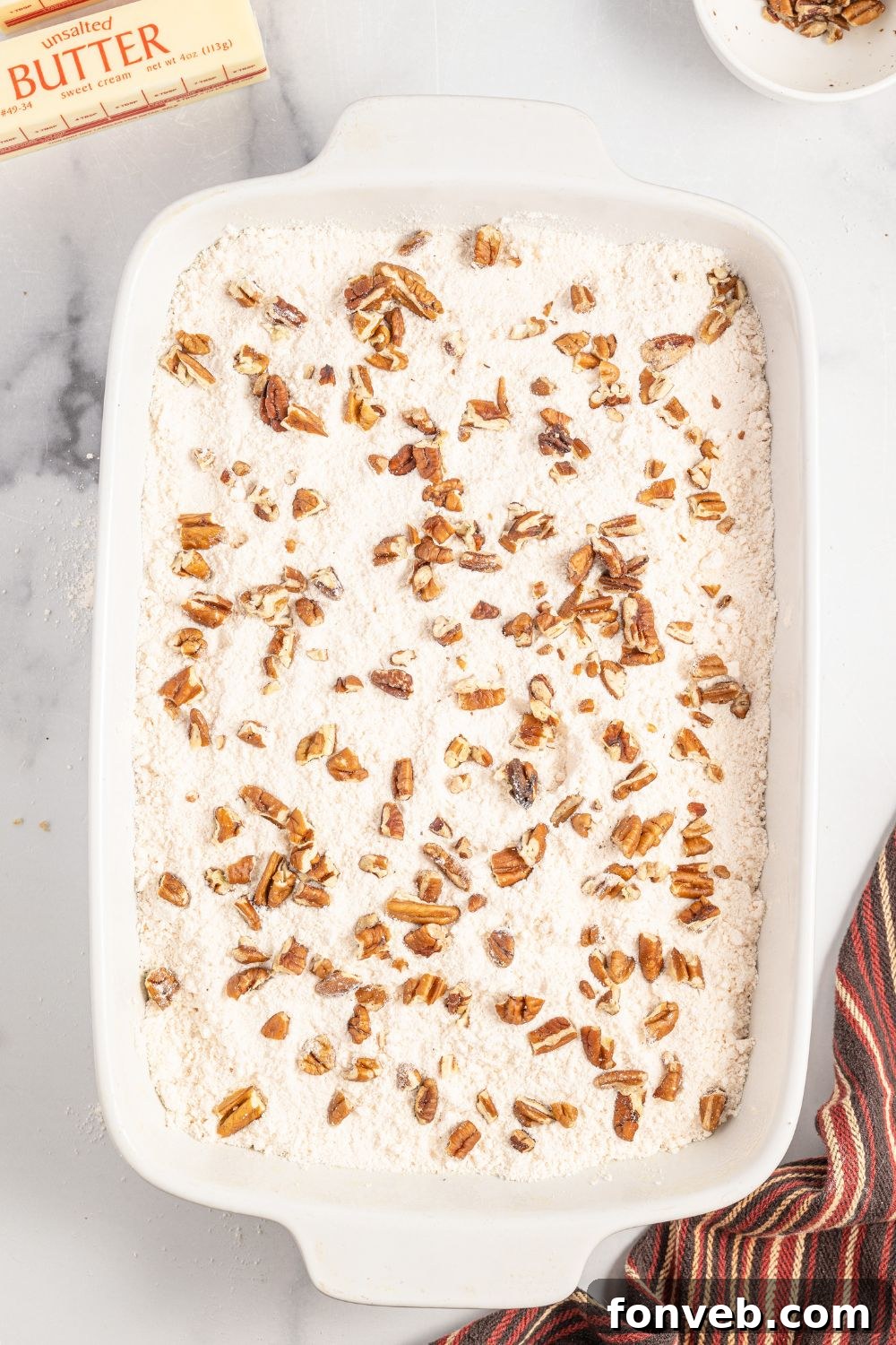 A layer of dry yellow cake mix evenly distributed over the pumpkin batter in a 9x13 baking dish, sprinkled with chopped pecans, awaiting the butter topping.