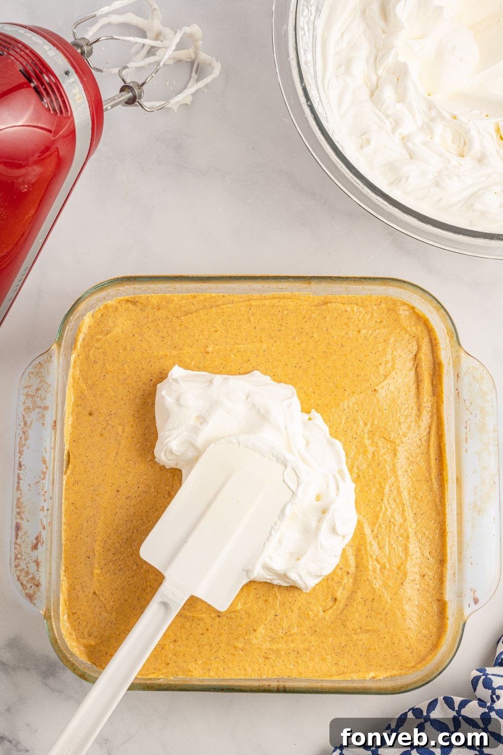 Homemade whipped cream being carefully spread over the top of the layered pumpkin dessert in the pan.