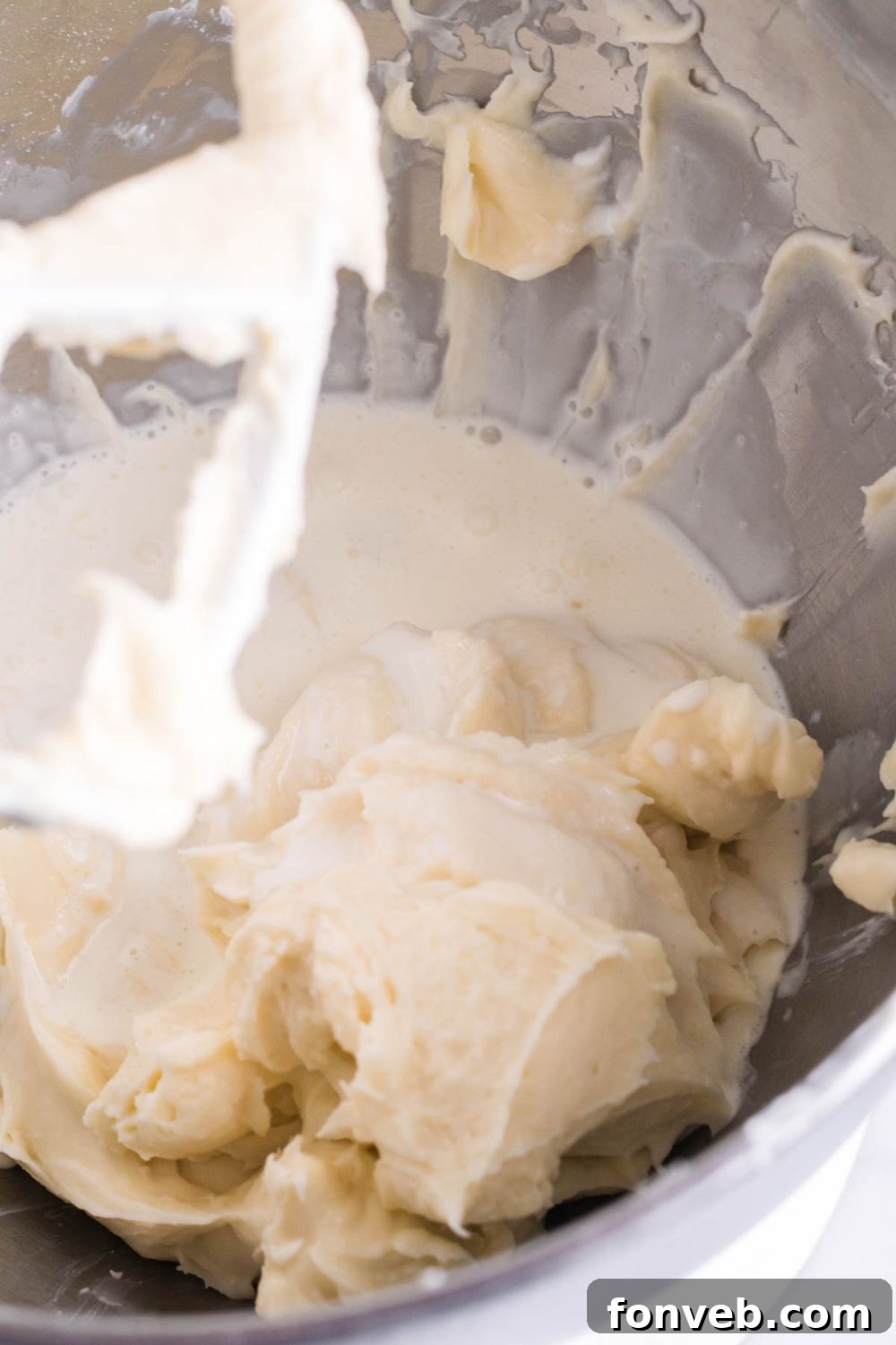 Close-up of a kitchen aid mixer bowl with a creamy white mixture, being beaten with a paddle attachment, indicating the preparation of the cream cheese filling.