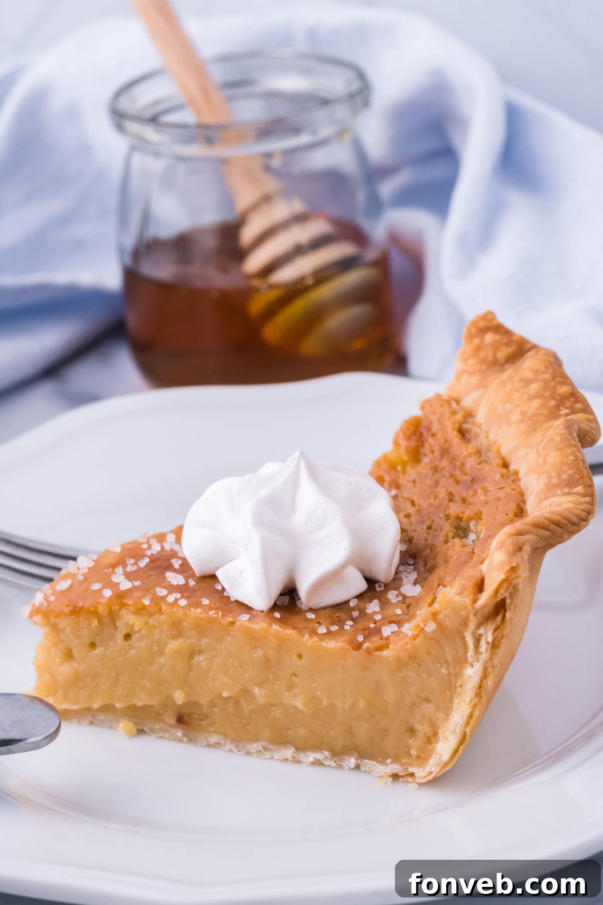 Golden Sweetheart 13 A close-up side view of a delectable honey pie slice on a white plate, with a clear jar of raw honey elegantly placed in the background.