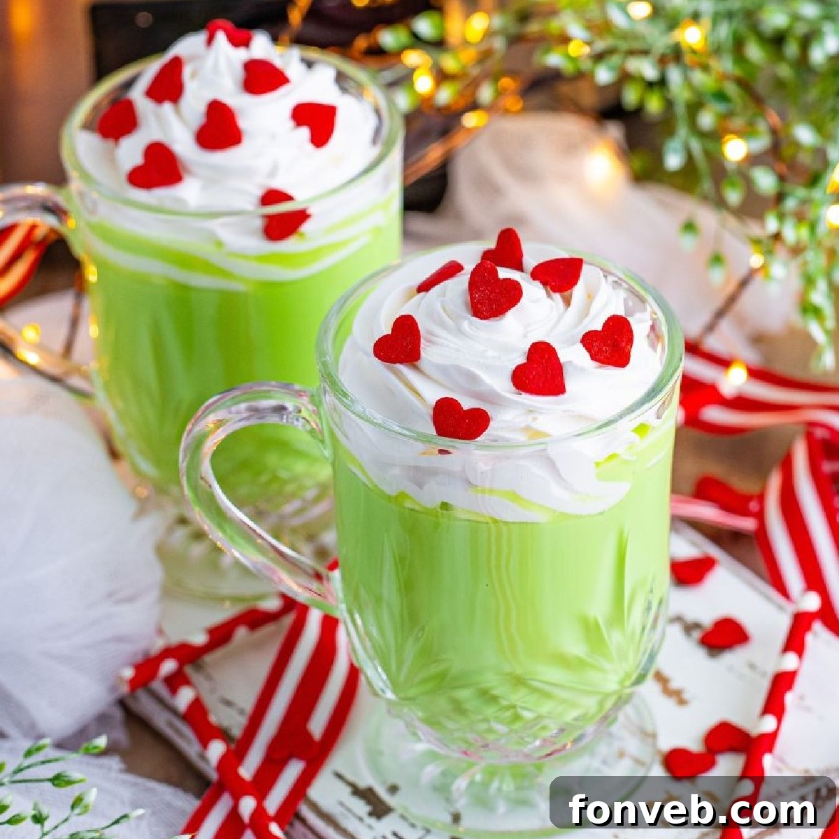 Grinch Hot Chocolate with whipped cream and red heart sprinkles in festive glasses on a white wood table