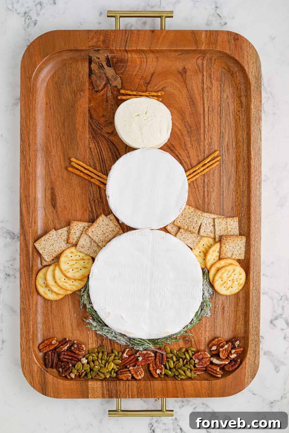 Brie cheese wheels arranged to form the body of a snowman on a serving tray, with other ingredients nearby