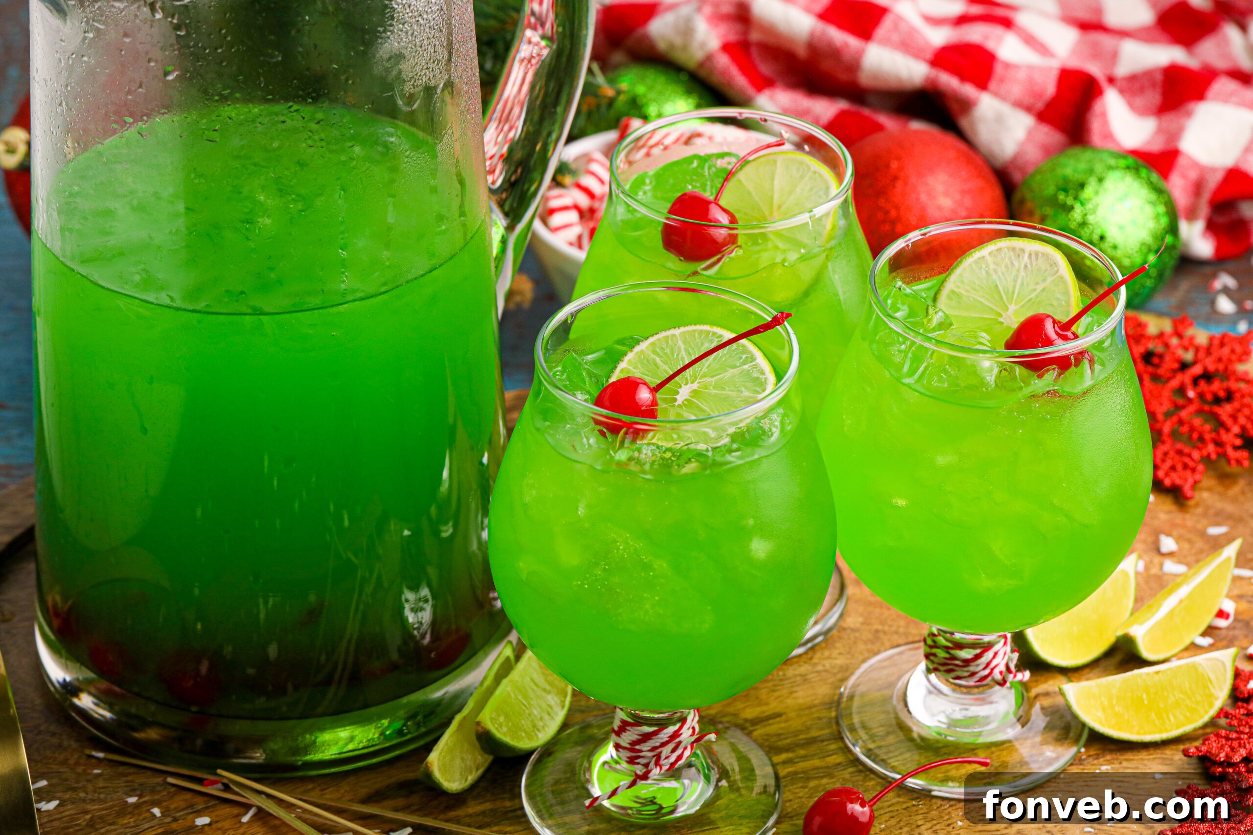 Grinch Punch in a clear glasses on a table with a cherry and a lime slice in the glasses, next to a large pitcher 