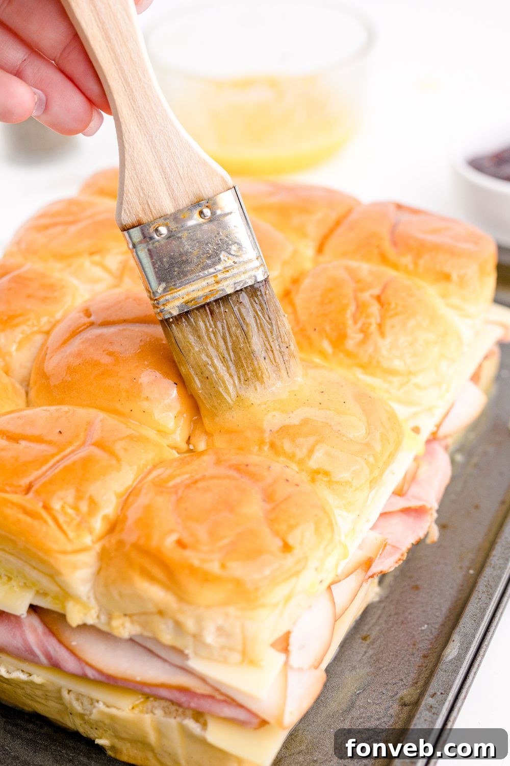 A hand brushing the tops of the slider buns with the butter and egg mixture before baking.