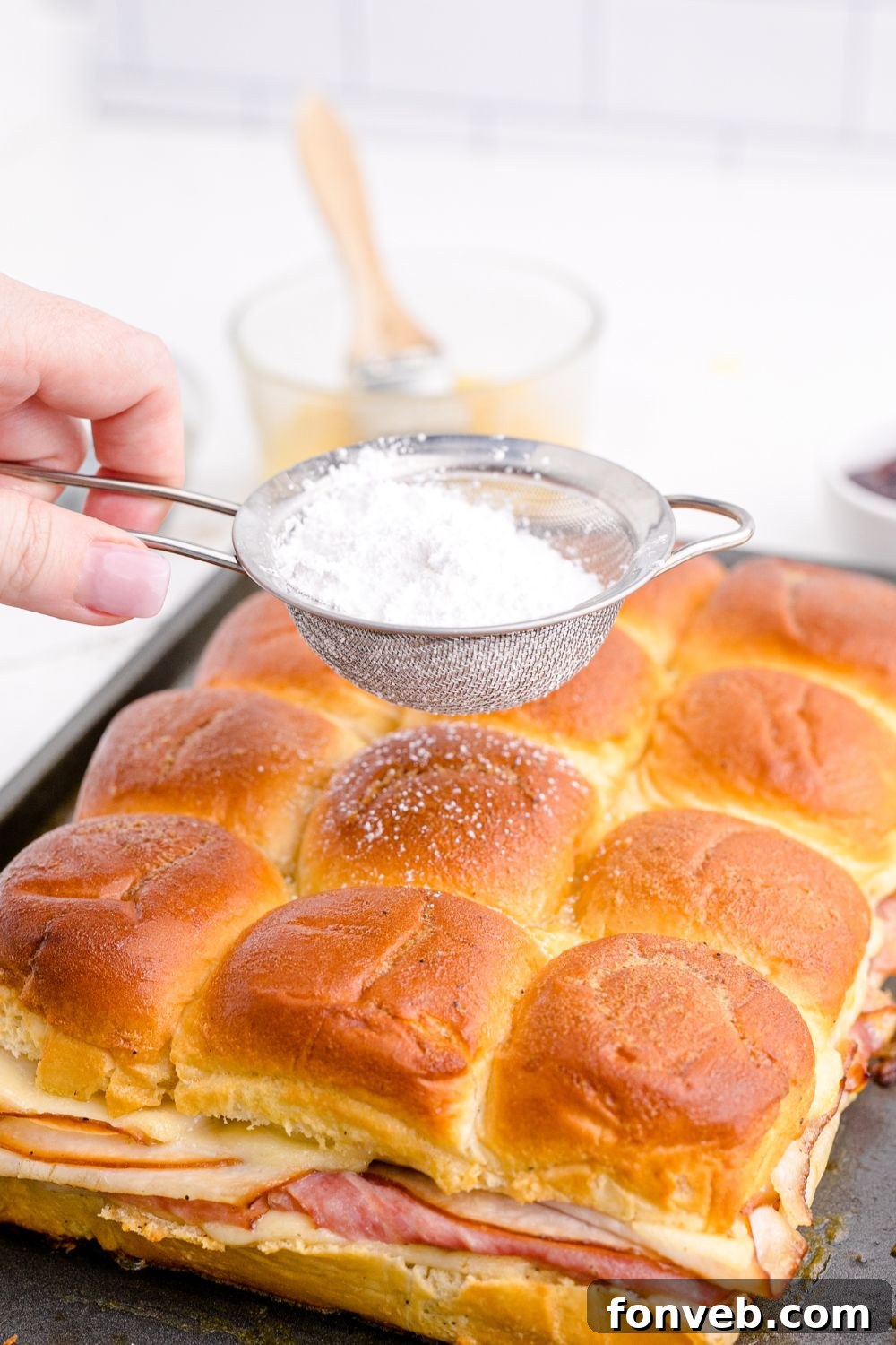 A mesh sieve being used to dust powdered sugar over the baked Monte Cristo Sliders.