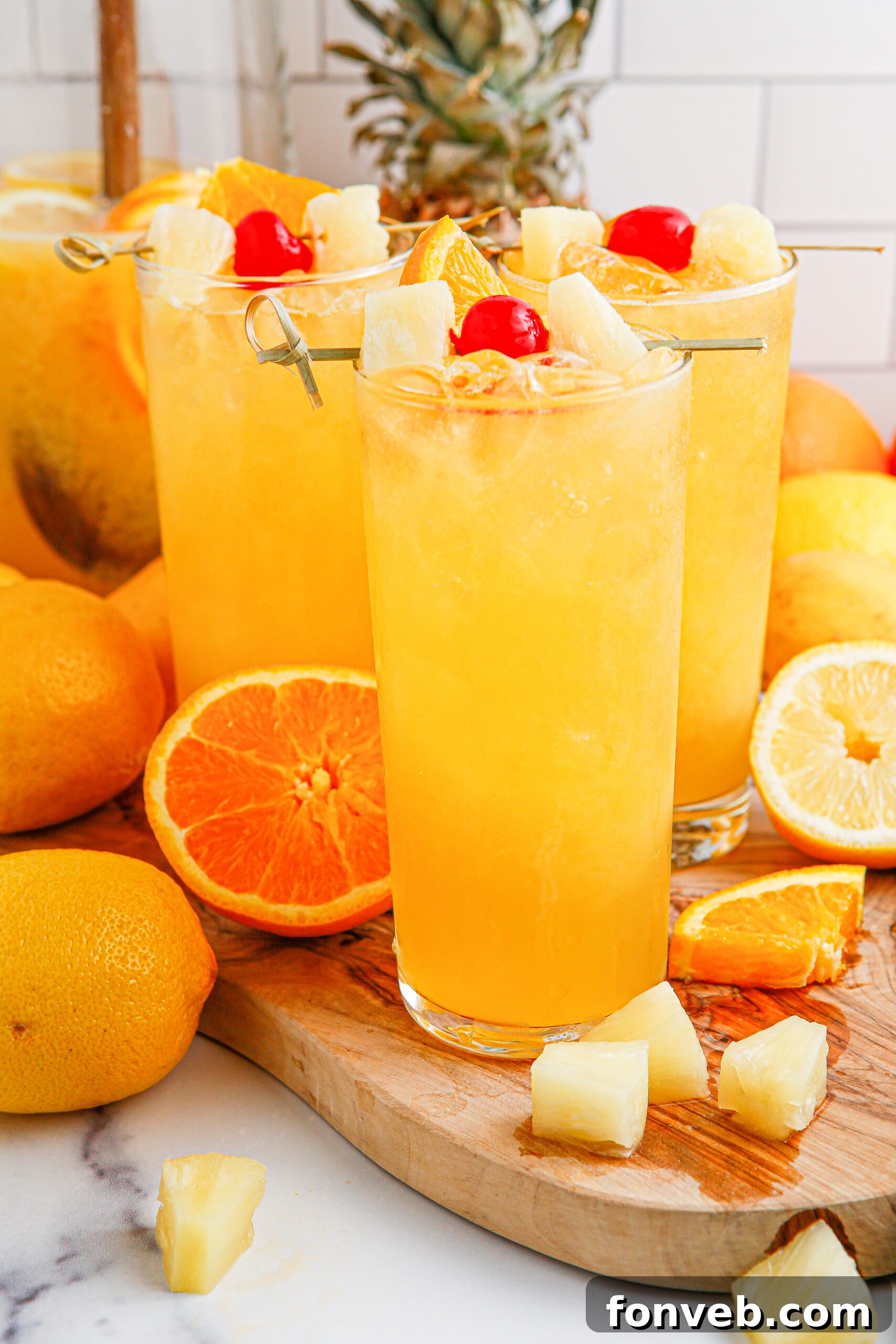 close up of the Tropical Lemonade Punch in 3 tall clear glasses on a wooden table with fruit around it and a pineapple in the background 