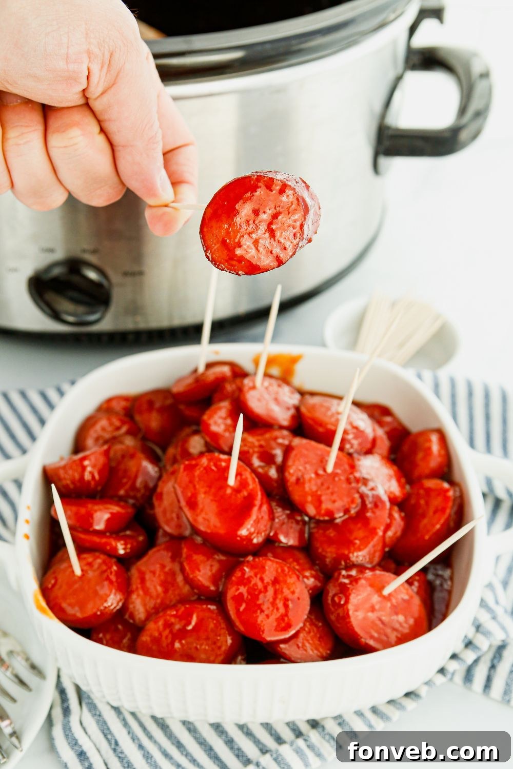 A person holding a toothpick with a perfectly glazed kielbasa bite, ready to eat.