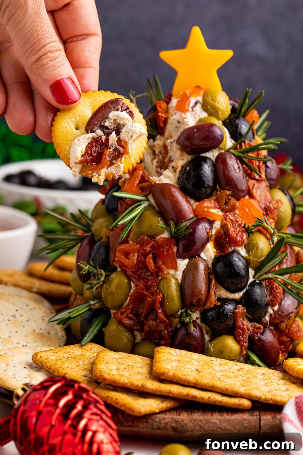 Cracker with cream cheese and olive dip on it by the cheeseball on table, showing a portion removed