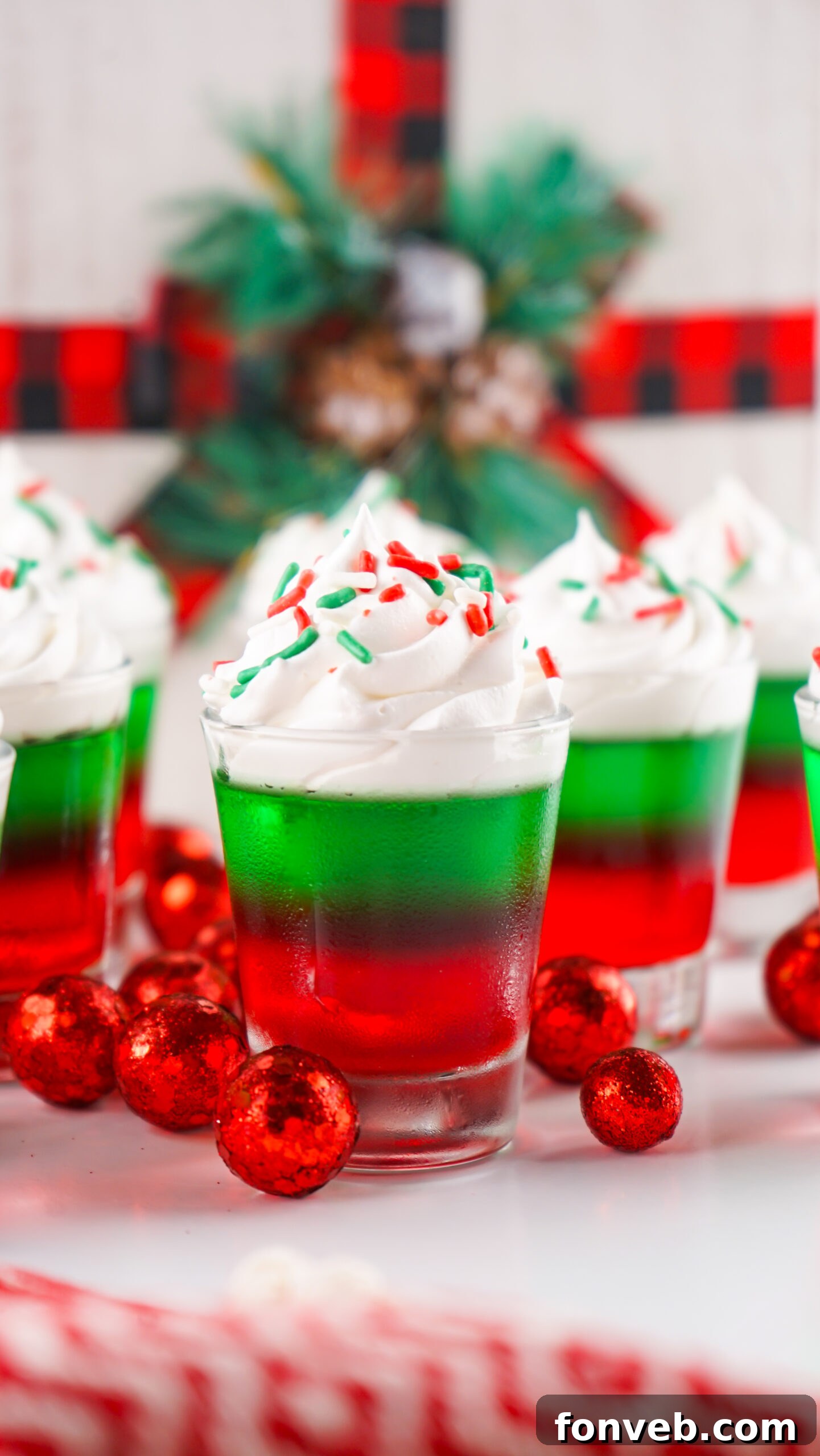 Yuletide Jiggle Shots 2 Christmas Jello Shots on a white able with red ornaments around them topped with holiday sprinkles and whipped cream