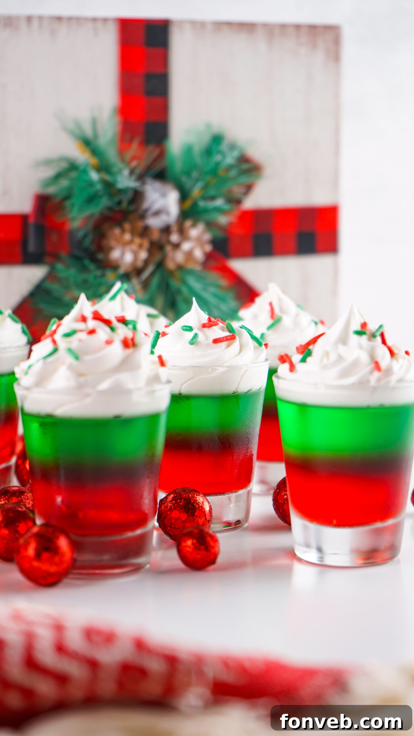 Yuletide Jiggle Shots 12 Christmas Jello Shots on a white able with red ornaments around them topped with holiday sprinkles and whipped cream