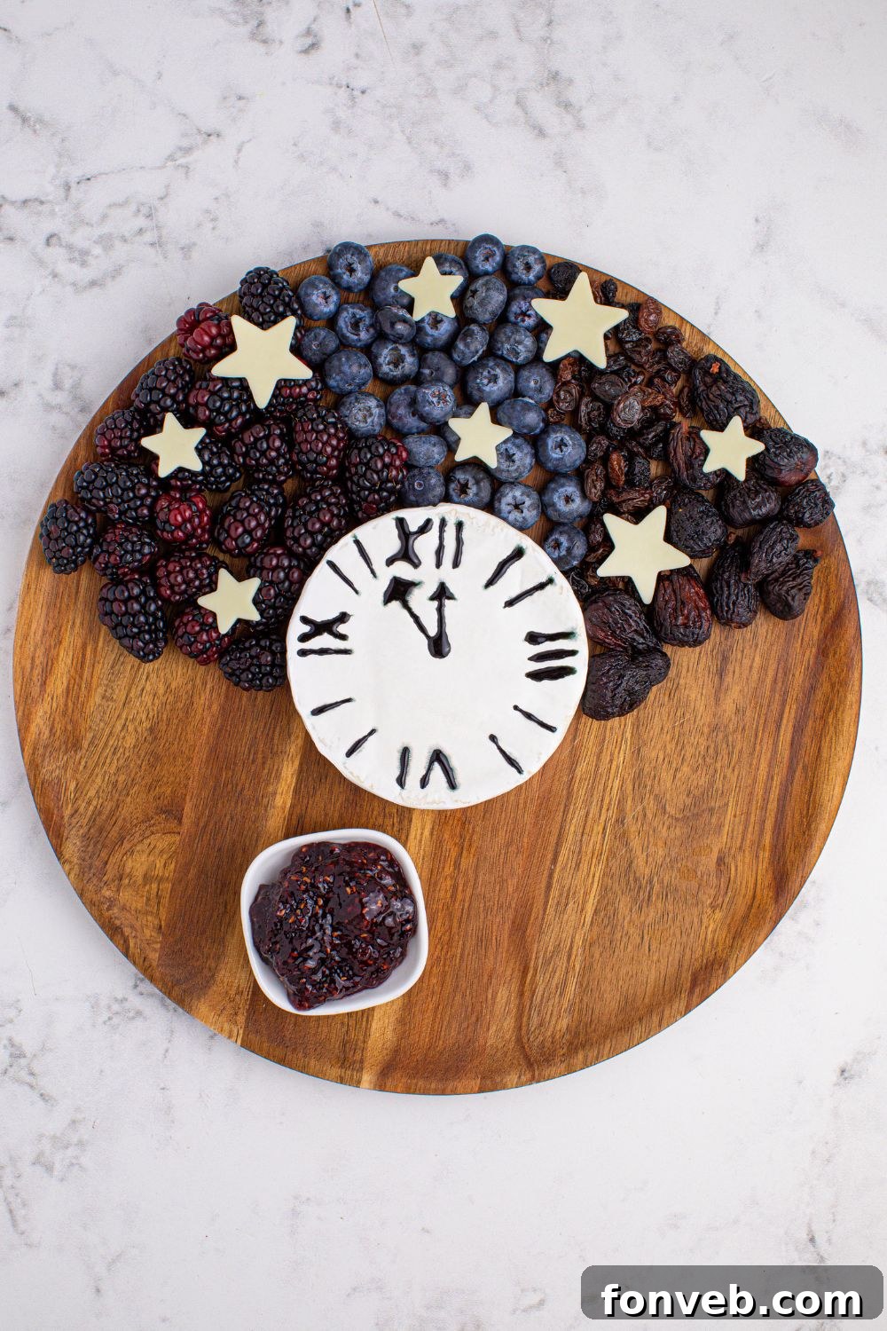 An assembly step for a charcuterie board, showing an arrangement of fresh berries and a Brie block, indicating how to begin organizing items on a round cutting board.