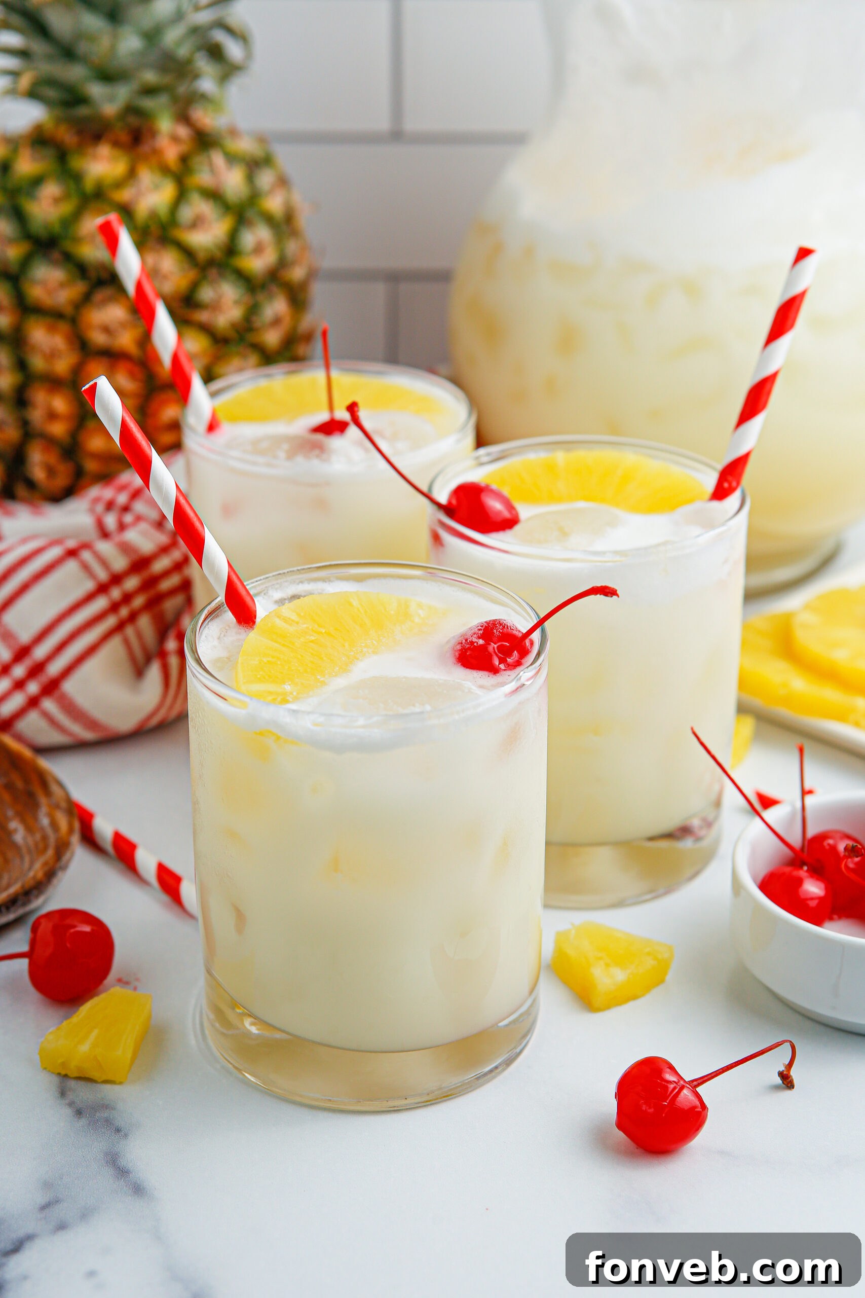 Island Escape Punch 2 Close-up of three glasses filled with frothy Pina Colada Punch, garnished with pineapple wedges and maraschino cherries, sitting on a wooden table.