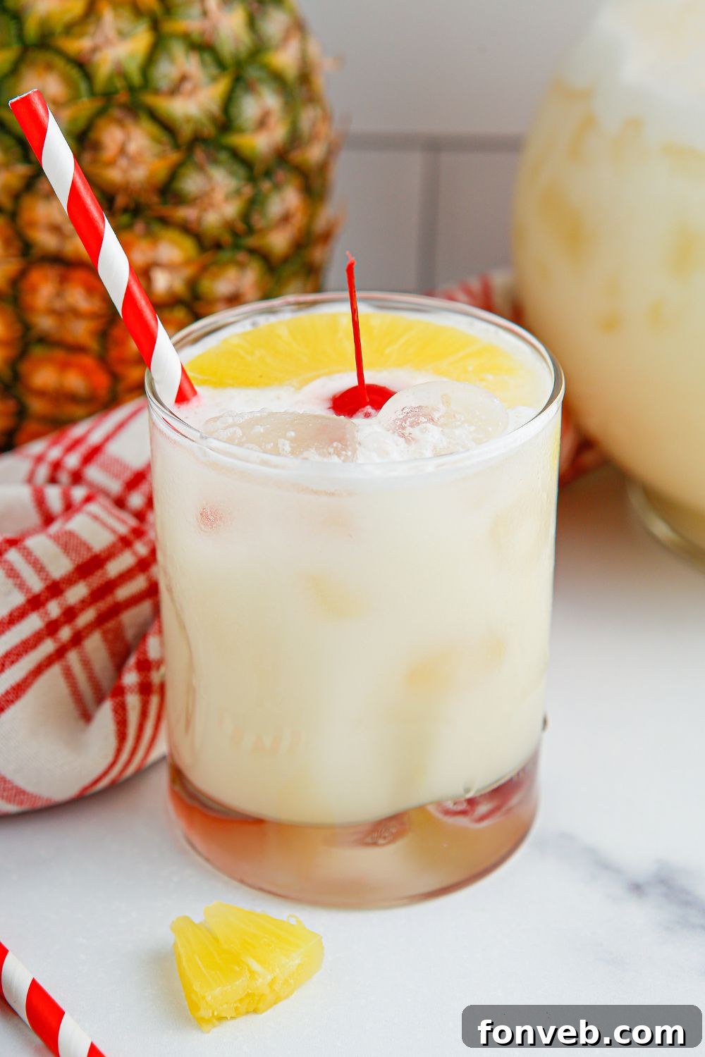 Island Escape Punch 11 A glass of Piña Colada Punch on a table, with a whole pineapple and a pitcher of punch blurred in the background.