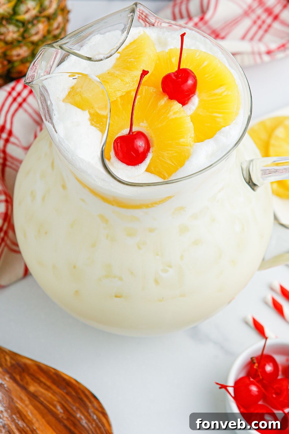 Island Escape Punch 13 A pitcher of Piña Colada Punch on a wooden table, surrounded by loose cherries, pineapple slices, and a wooden stirring spoon.