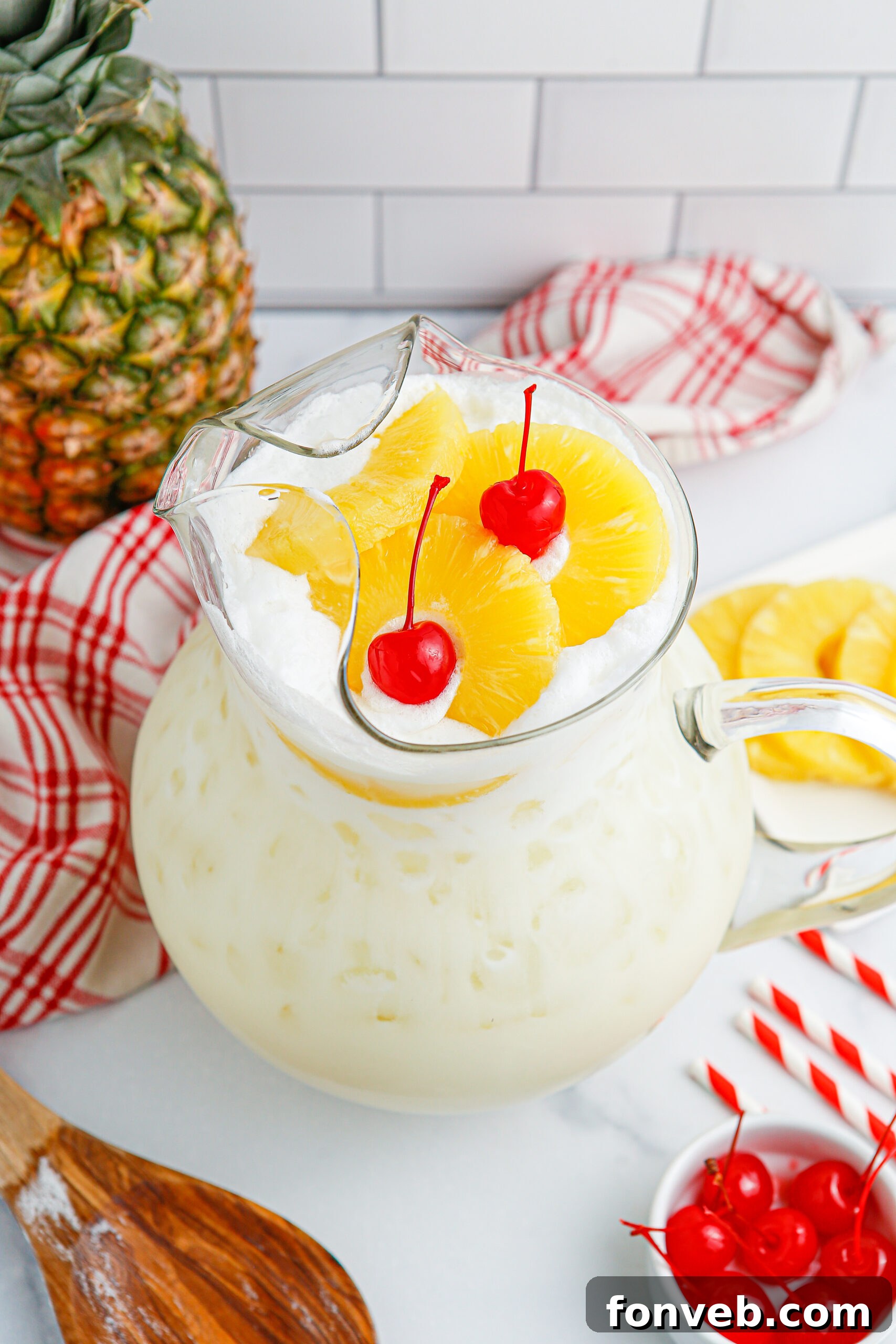 Island Escape Punch 15 A pitcher of Piña Colada Punch on a wooden table with pineapple slices and cherries, next to a wooden spoon.