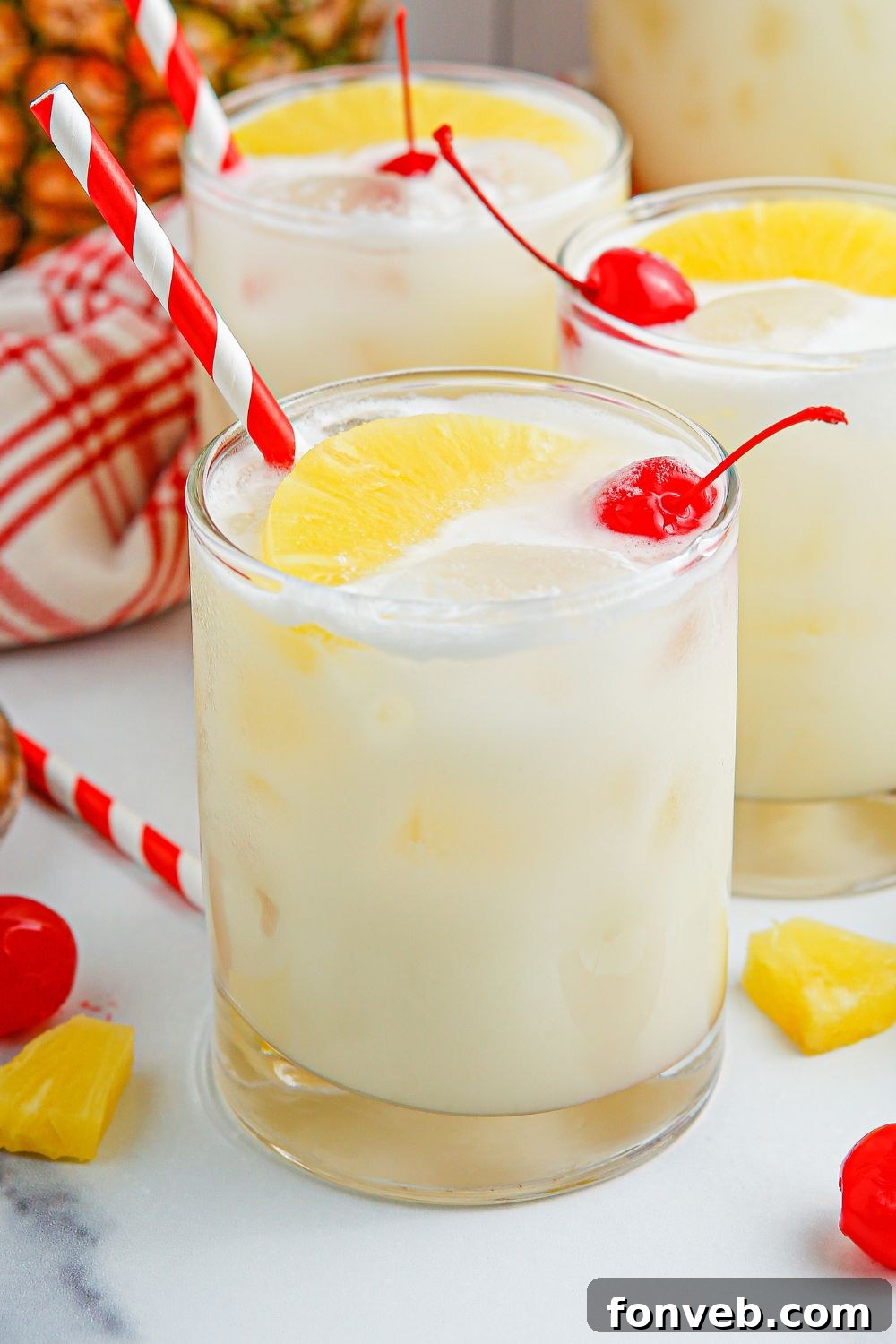 Island Escape Punch 18 Three glasses of Piña Colada Punch on a table, garnished with pineapple and cherries, surrounded by tropical decor.