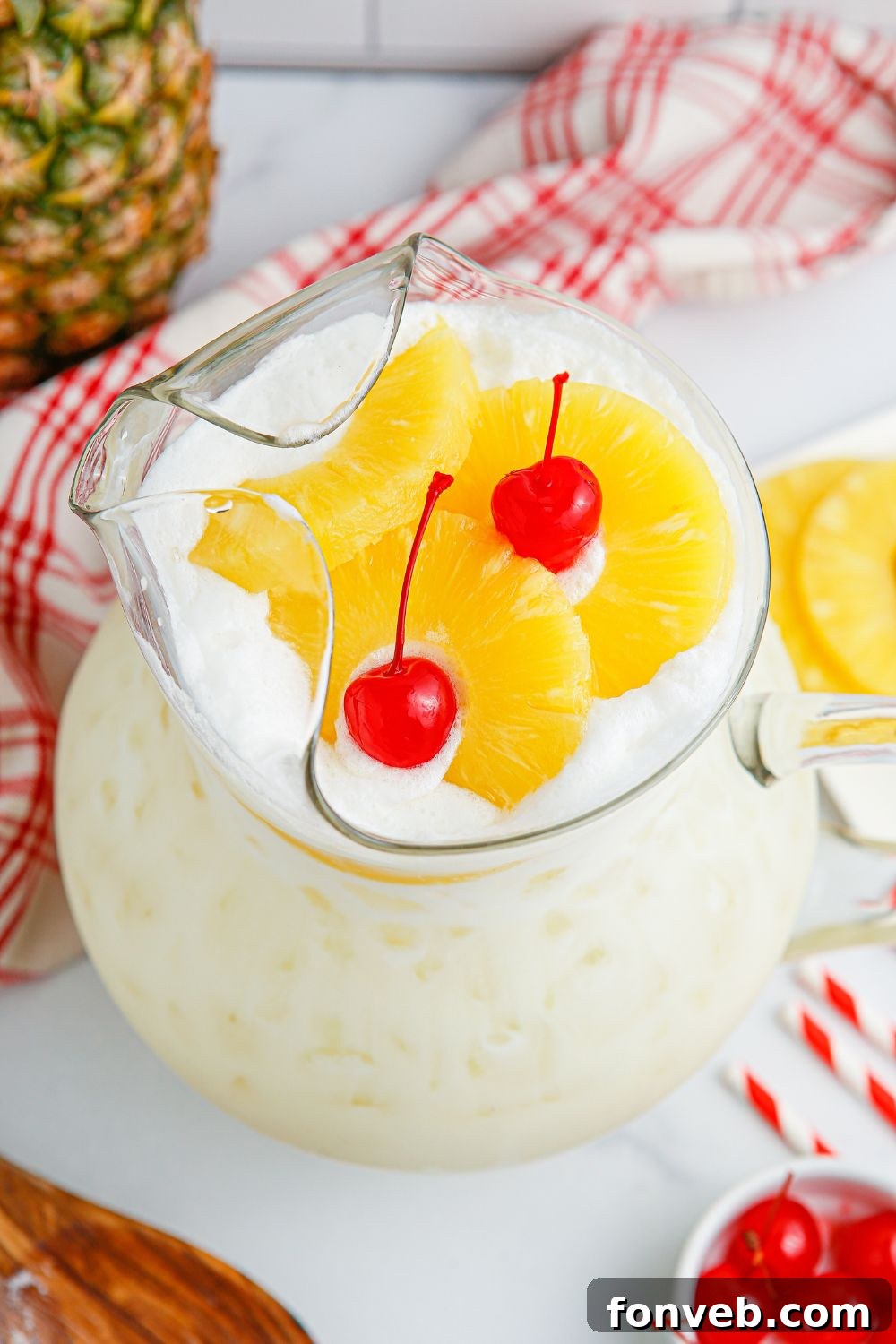 Island Escape Punch 4 A pitcher of Piña Colada Punch on a table, beautifully garnished with fresh pineapple slices and bright red cherries floating on top.