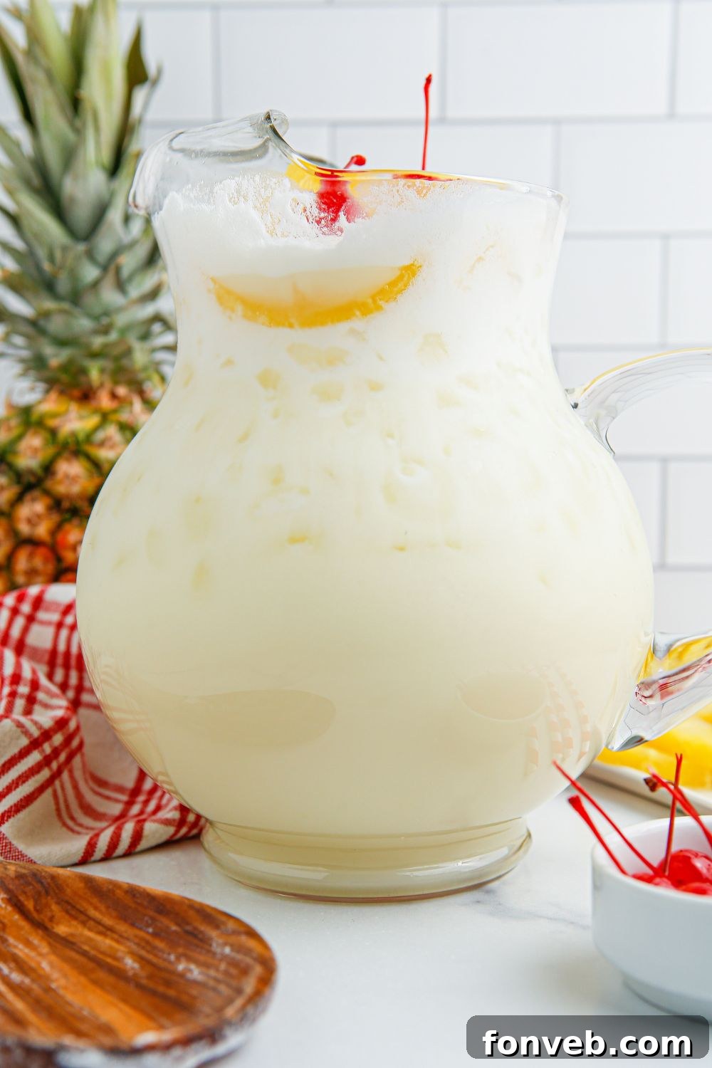 Island Escape Punch 8 A large pitcher of Piña Colada Punch on a table, accompanied by a whole pineapple and a bowl of maraschino cherries, ready for garnishing.
