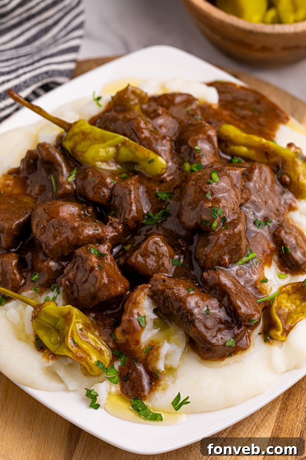 Tender Mississippi Crockpot Steak Bites 3 A plate of Mississippi Steak Bites with mashed potatoes underneath, a side of pepperoncinis, and a slow cooker in the background.