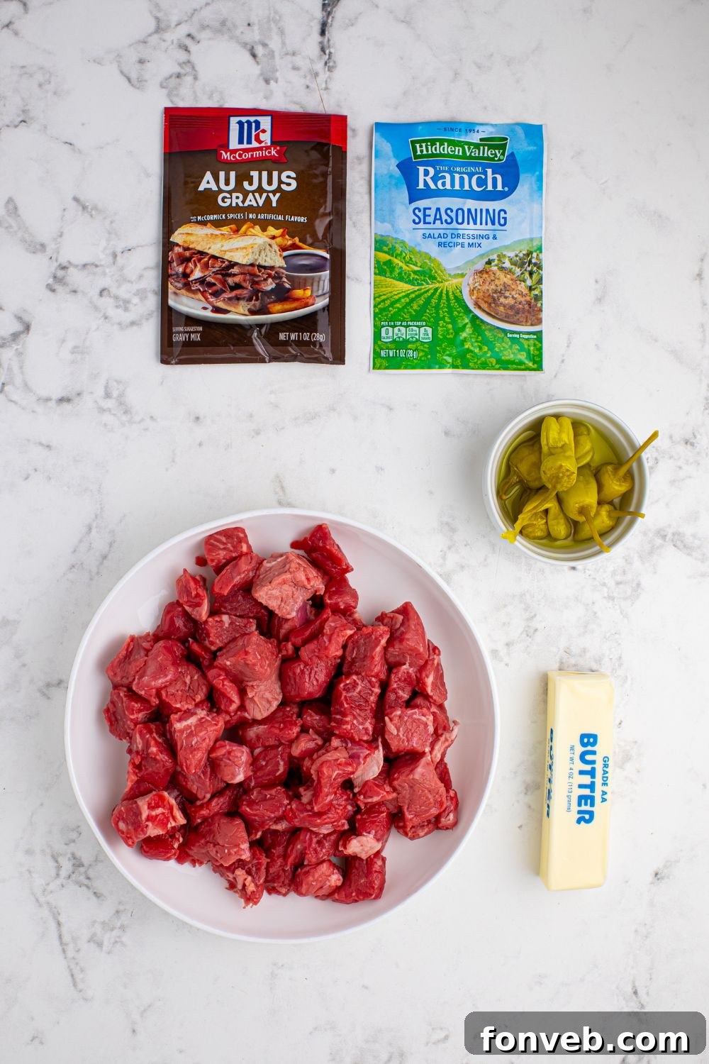 Tender Mississippi Crockpot Steak Bites 6 Key ingredients for Mississippi Steak Bites laid out: steak cubes, ranch seasoning, au jus gravy mix, butter, and pepperoncini peppers.