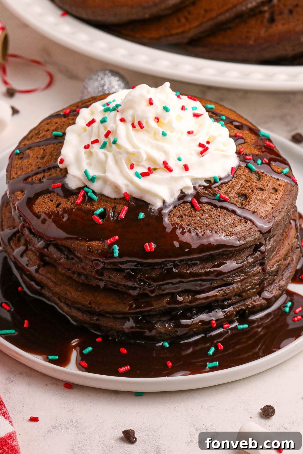 Decadent Hot Chocolate Pancakes 20 Hot Chocolate Pancakes stacked on a plate with hot chocolate and whipped cream and festive sprinkles