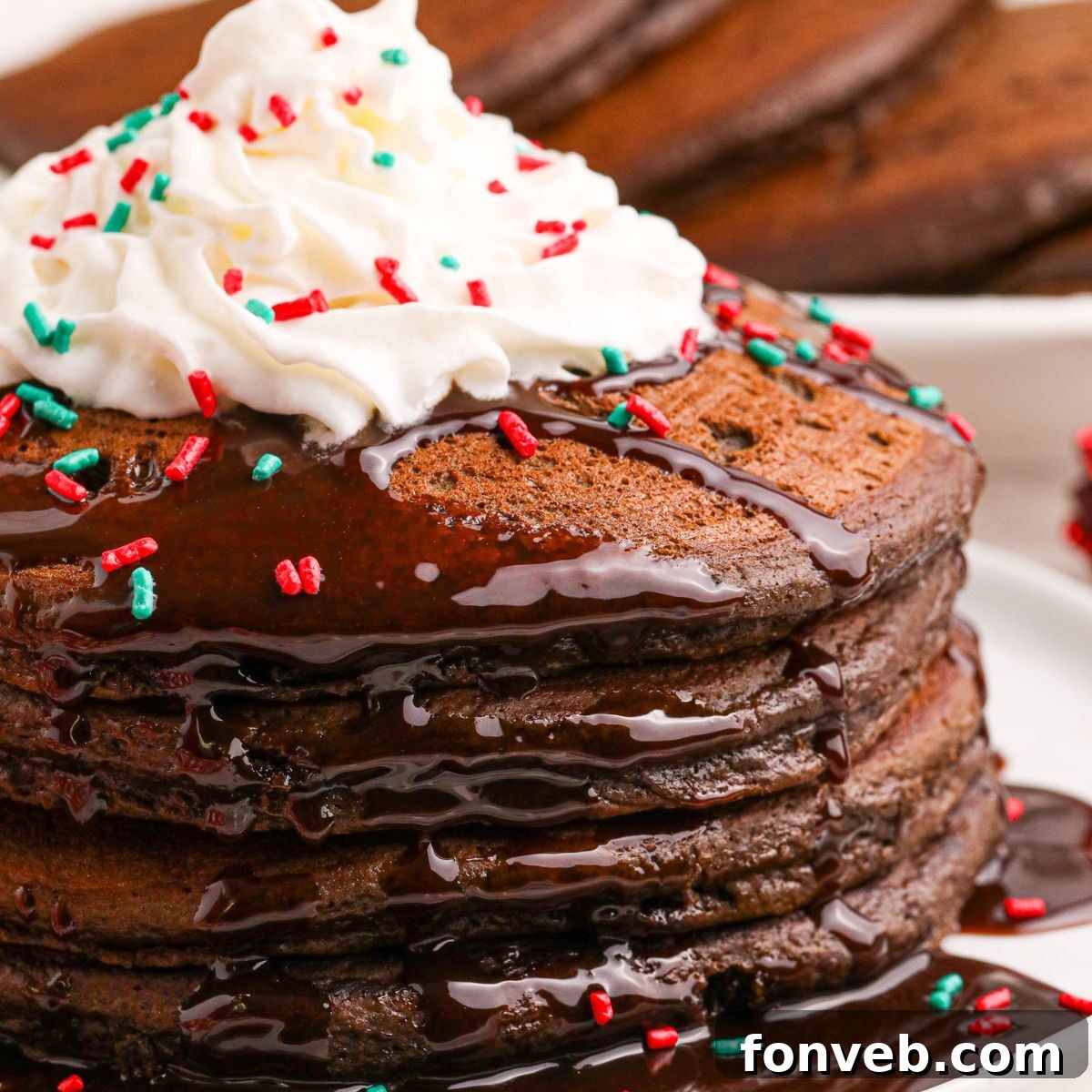 Decadent Hot Chocolate Pancakes 3 Hot Chocolate Pancakes stacked on a plate topped with syrup and whipped cream and holiday sprinkles