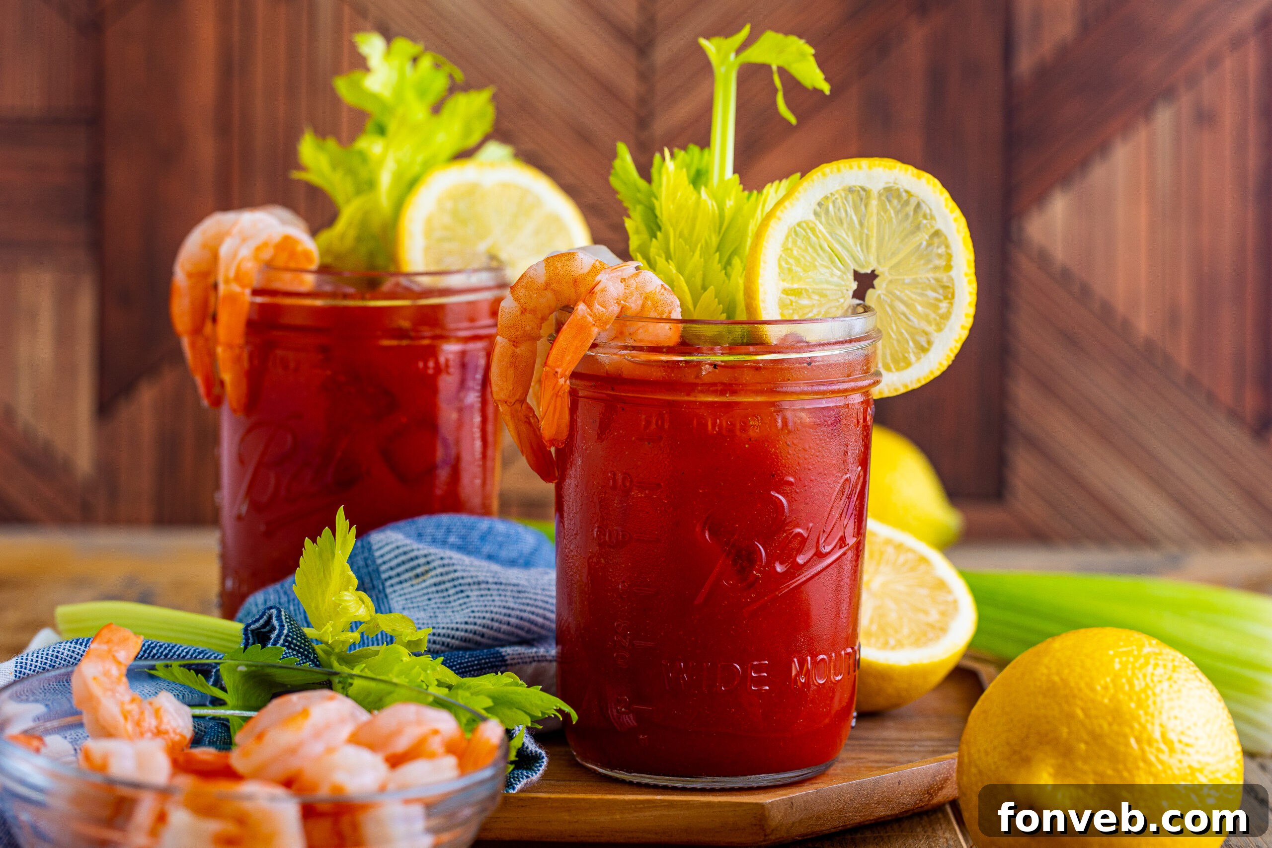 Bloody Mary in a clear mason jar glass topped with celery, sliced lemon and shrimp on a wooden table.